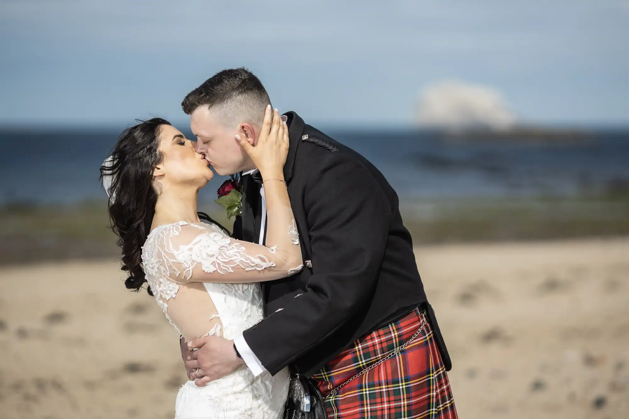A bride and groom kissing on a beach, with the groom wearing a kilt. A distant cloud drifts over the sea in the background.