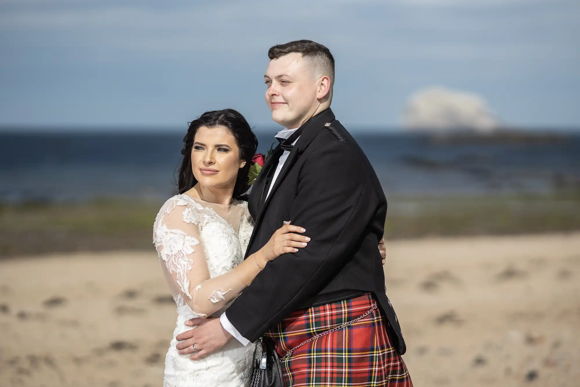 A bride wearing a white lace dress and a groom in a kilt embrace on a sandy beach with the ocean and a cloudy sky in the background.