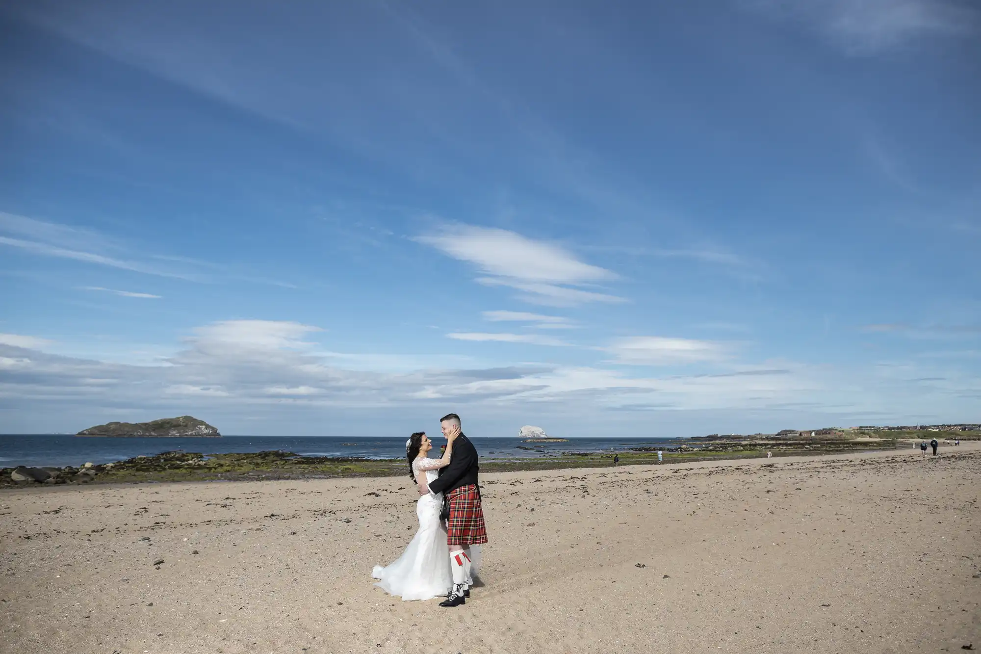 A couple in wedding attire, the man in a kilt, embracing on a sandy beach with distant islands under a clear sky.