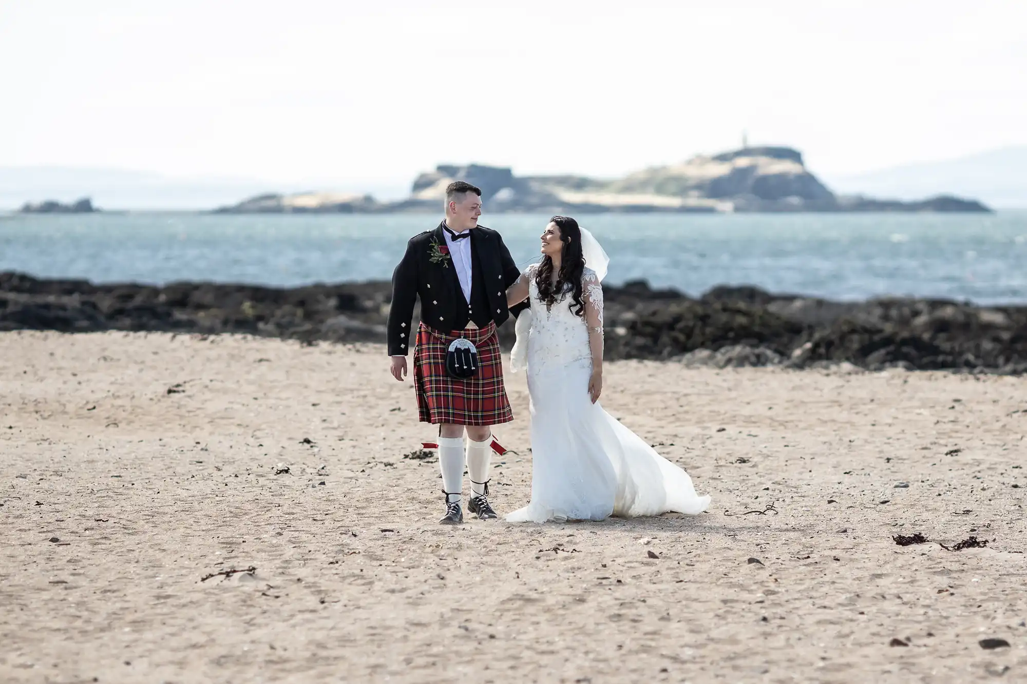 Bride in a white dress and groom in a Scottish kilt walking on a sandy beach with an island in the background.