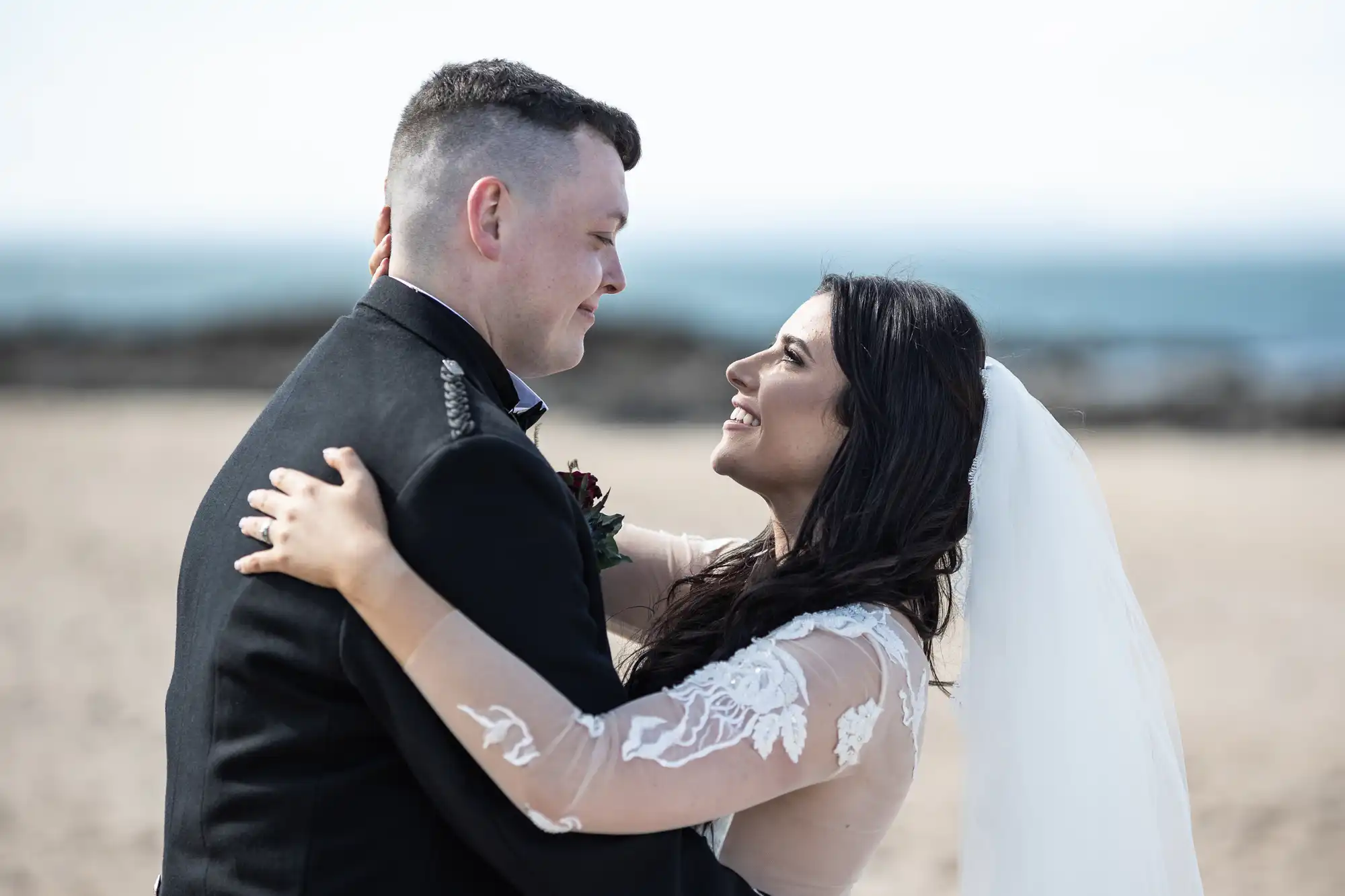 A bride in a lace dress and a groom in a black kilt share a joyful embrace on a sandy beach under a clear sky.