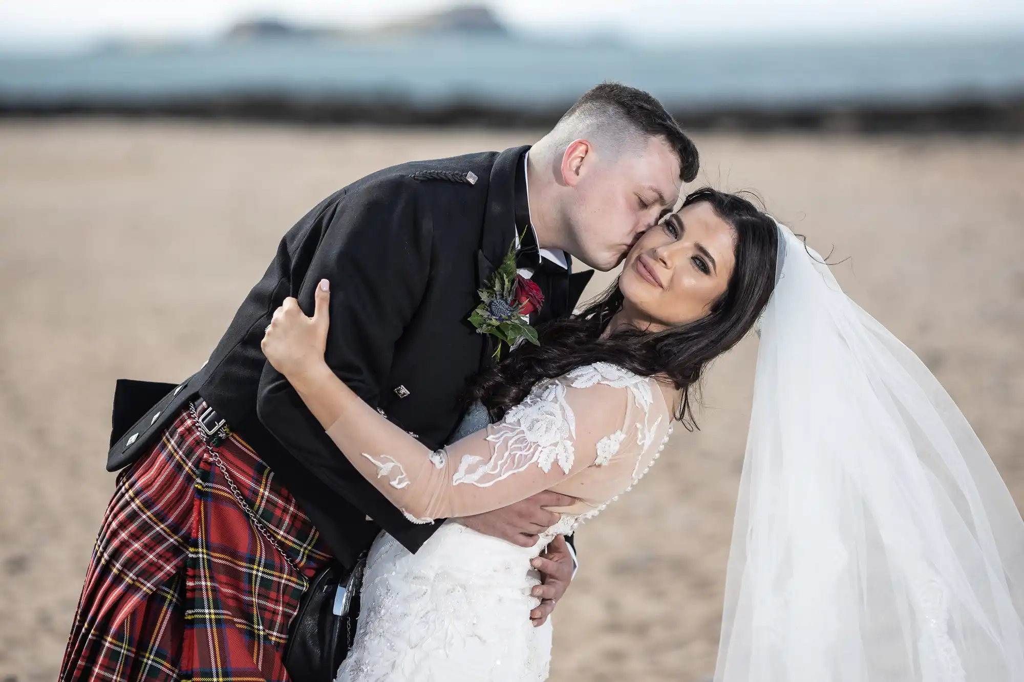 A groom in a kilt kissing a bride in a white lace dress on a beach.