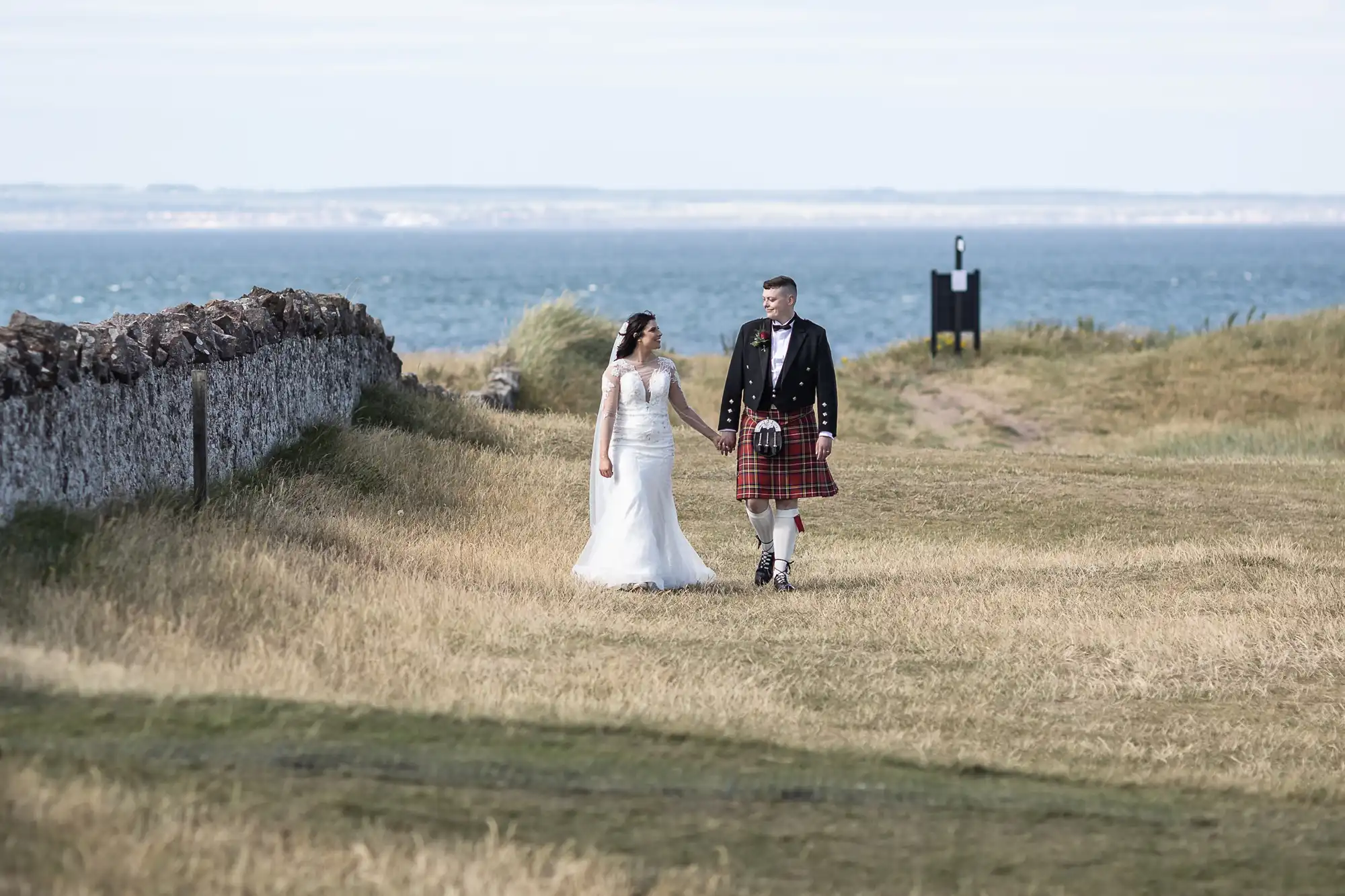 Bride in white dress and groom in traditional Scottish kilt holding hands on a coastal path with a stone wall and sea in the background.