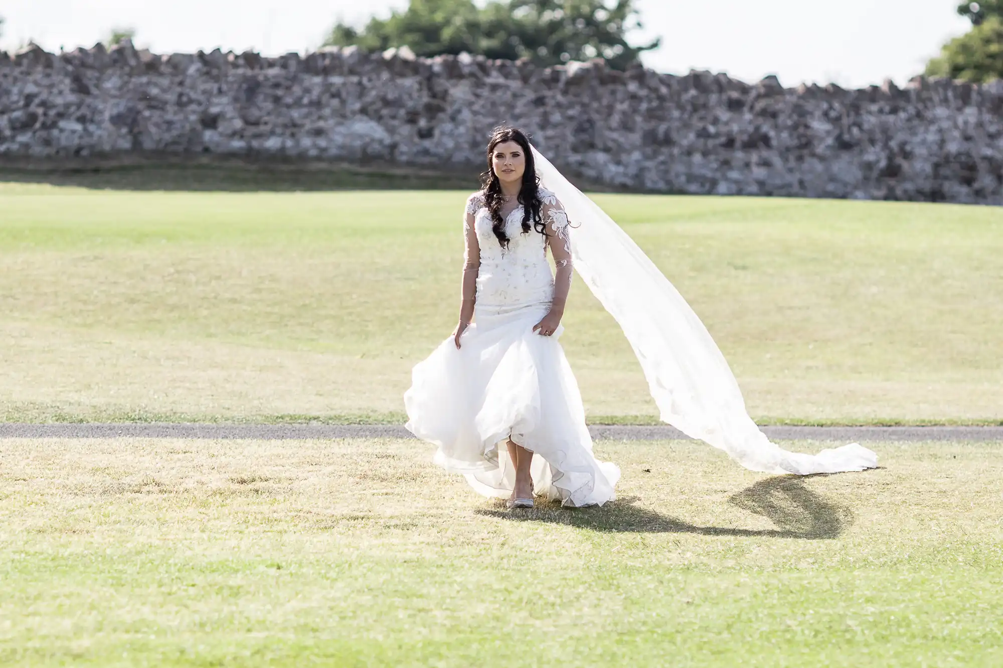 A bride in a white dress with a long veil walks across a grassy field, with an old stone wall in the background.
