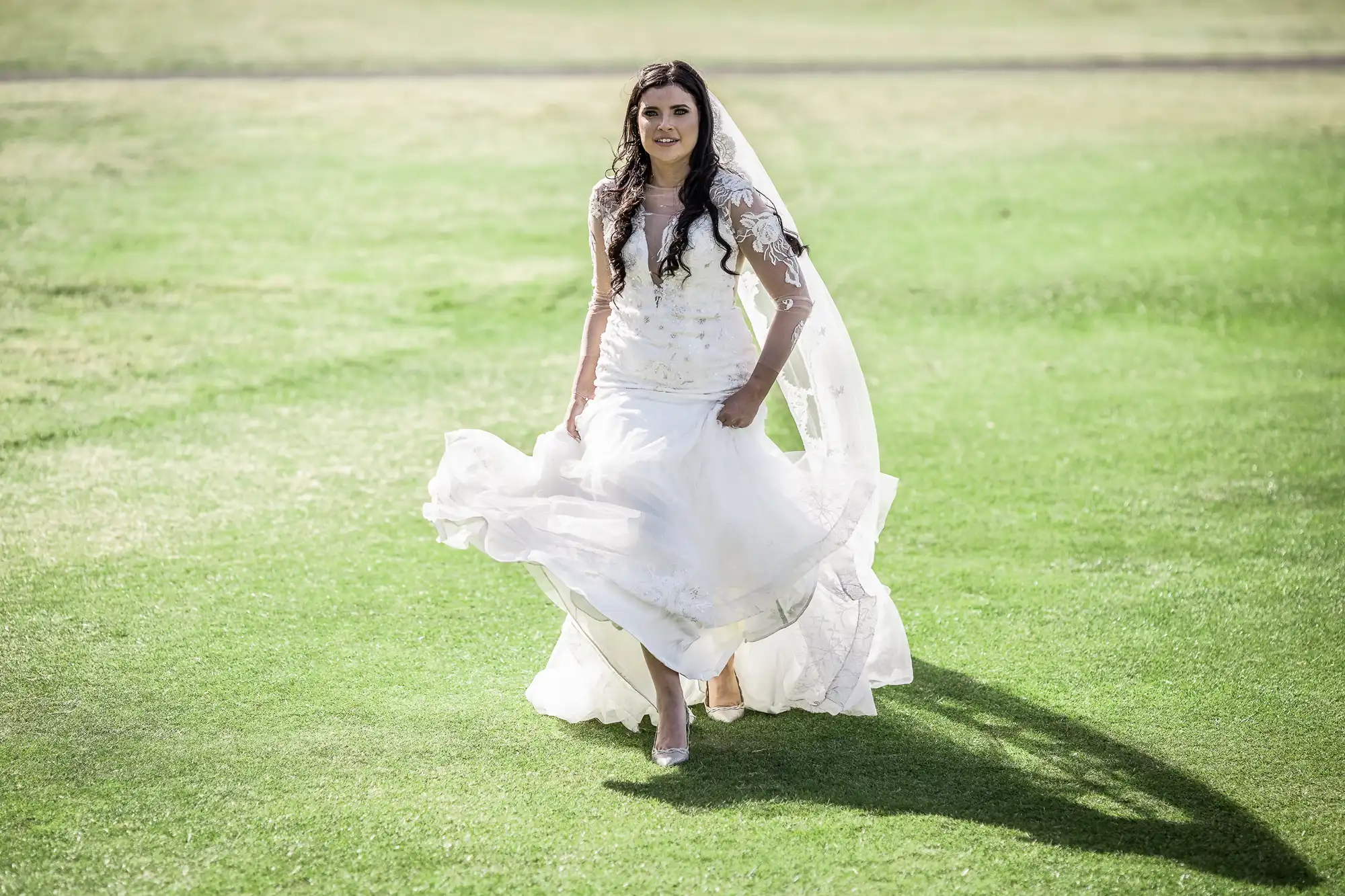 A bride in a flowing white dress walking on a grassy field, her dress and hair moving gently with her stride.