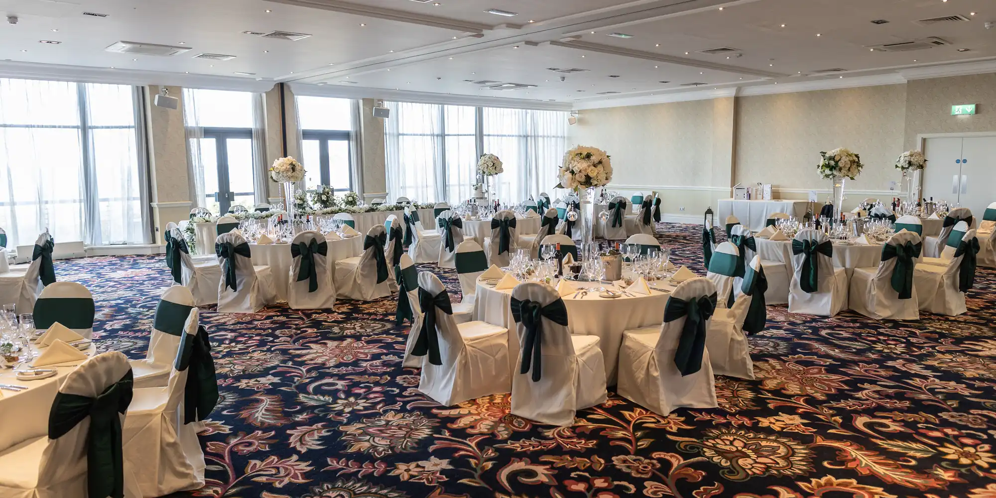 Elegant banquet hall setup with round tables, white linens, dark green chair sashes, and floral centerpieces, under soft lighting.