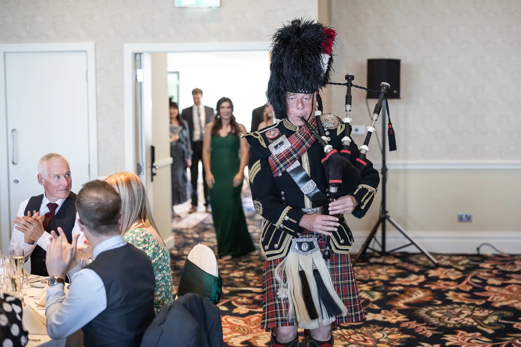 A man in traditional Scottish attire playing bagpipes at an indoor event, with guests seated at tables looking on.