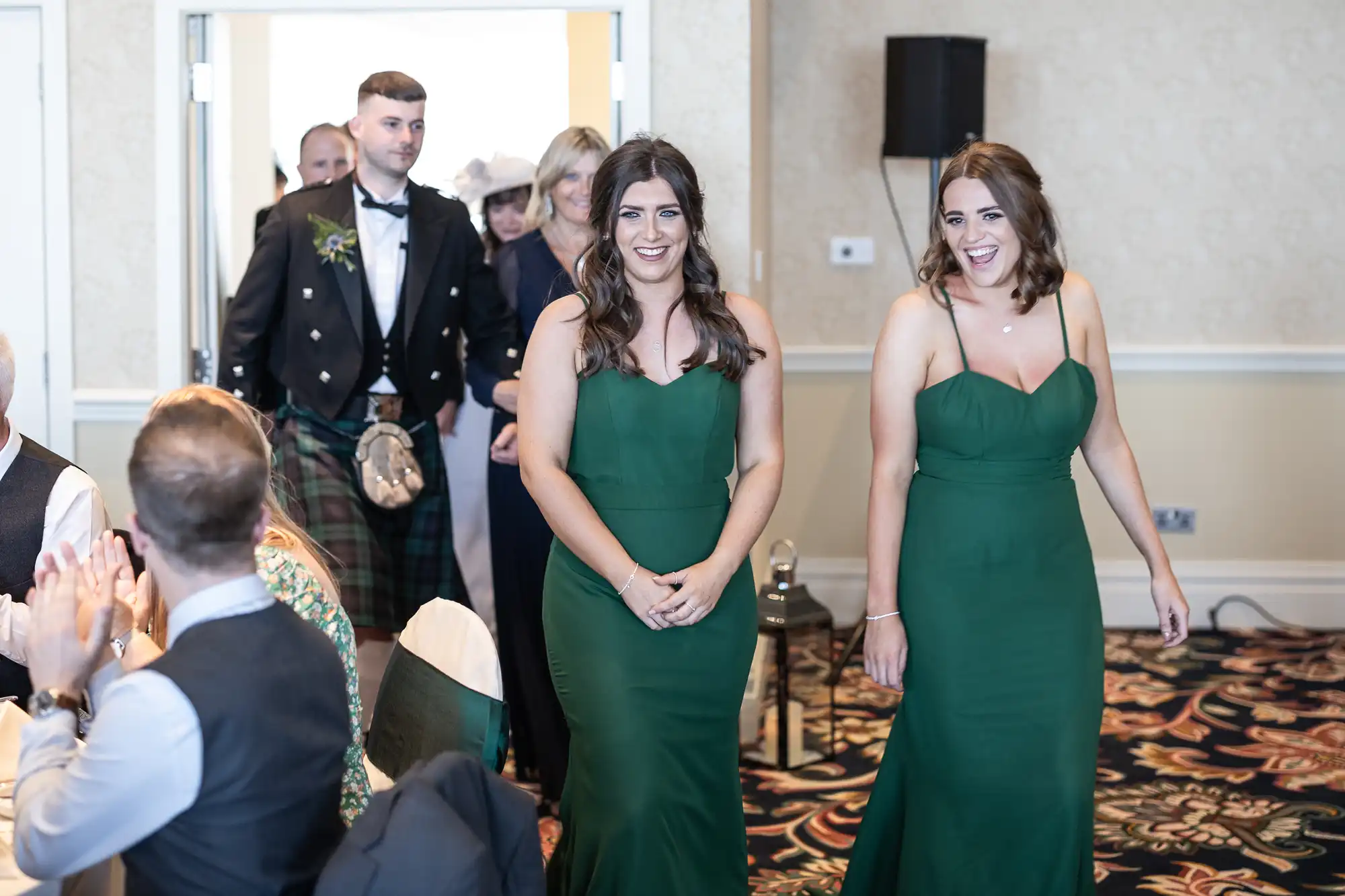 Two women in green dresses smiling and walking through a room of applauding guests at a formal event.