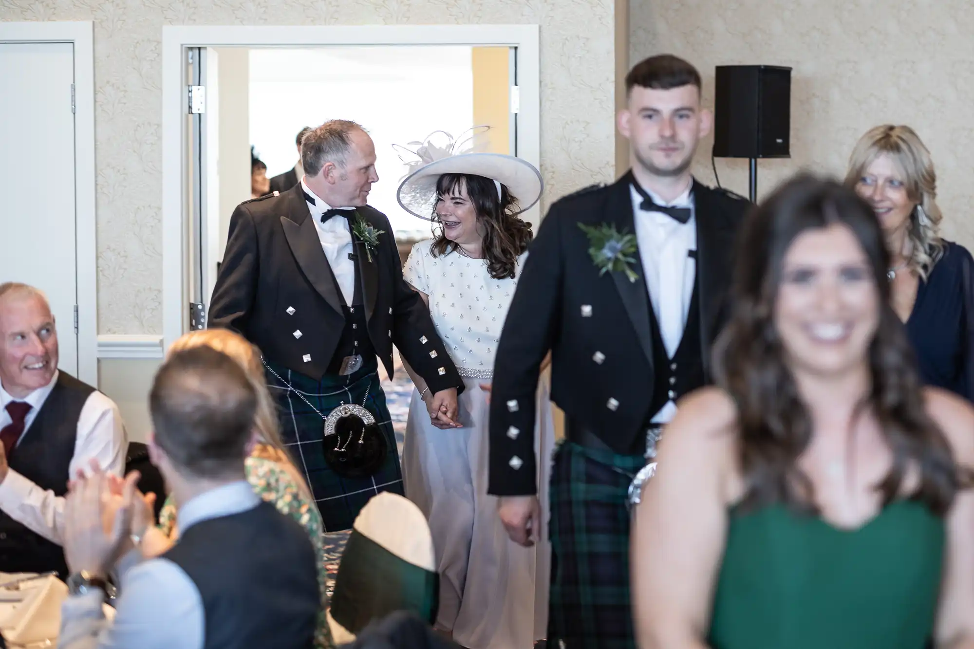A couple in wedding attire, the man in a kilt, enters a reception room greeted by applauding guests, with other sharply dressed attendees in the foreground.