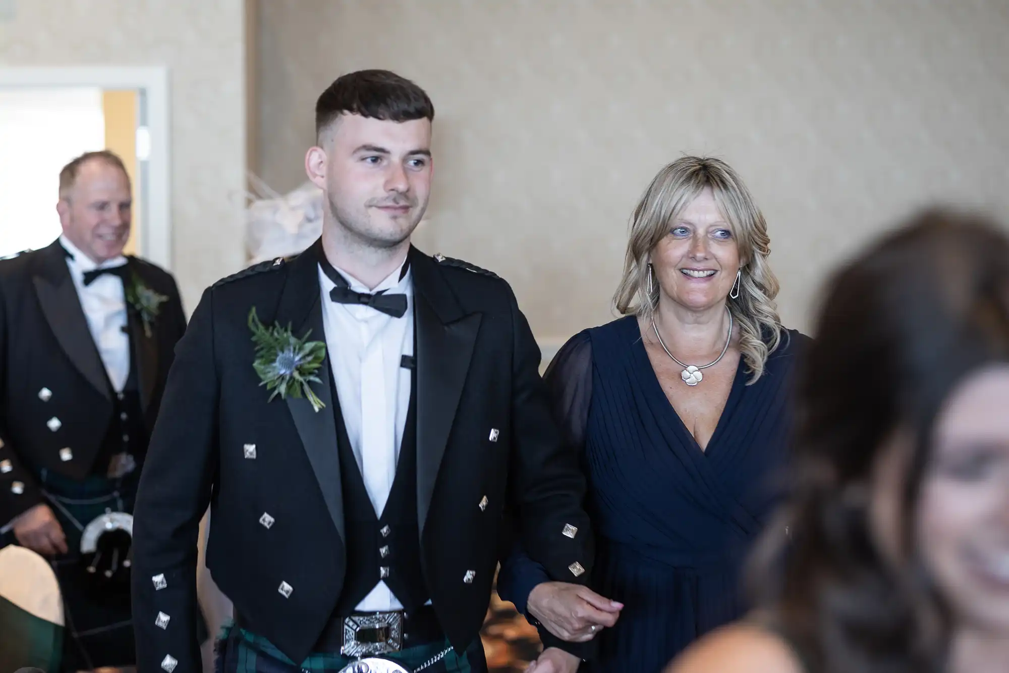 A young man in a kilt and a middle-aged woman walk together smiling in a crowded room during a festive event.