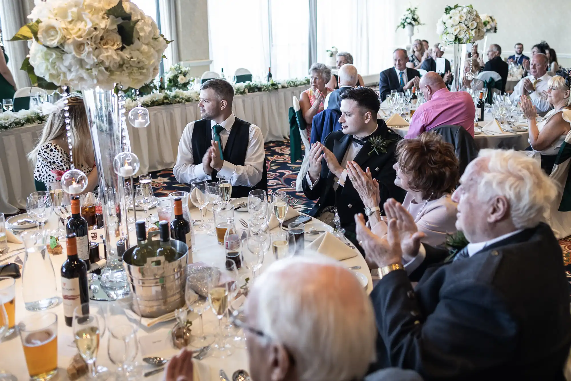 Guests at a wedding reception clapping, with two men at the foreground looking at each other, surrounded by tables adorned with floral decorations and wine.