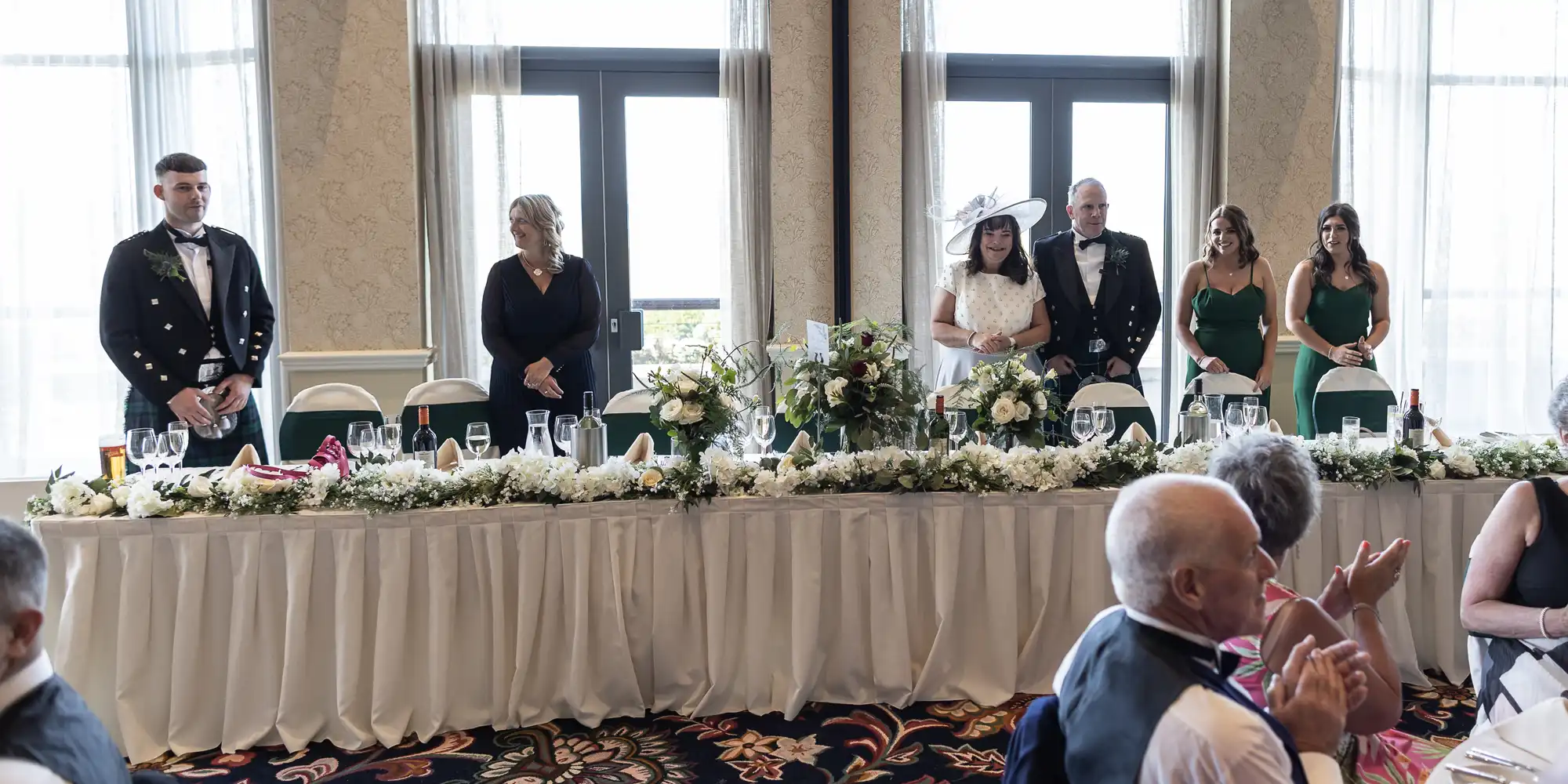 A wedding head table with a bridal party standing as guests applaud, featuring elegant floral decorations.