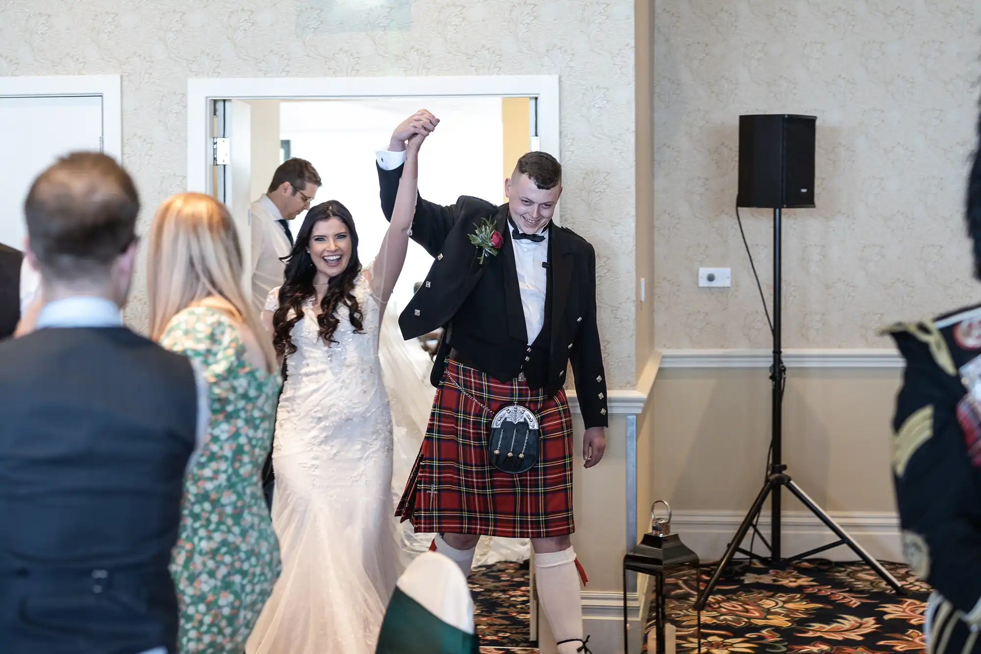 A bride and groom joyfully enter a reception hall, the groom wearing a traditional Scottish kilt, as guests applaud.