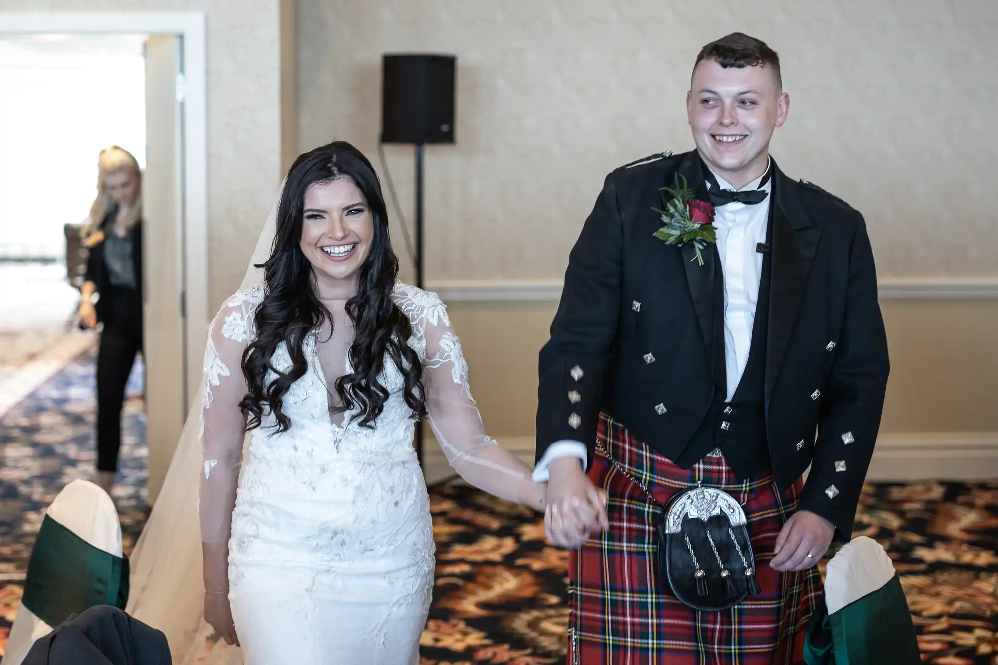 A bride in a lace dress and a groom in a traditional kilt smiling as they walk hand in hand through a wedding venue.