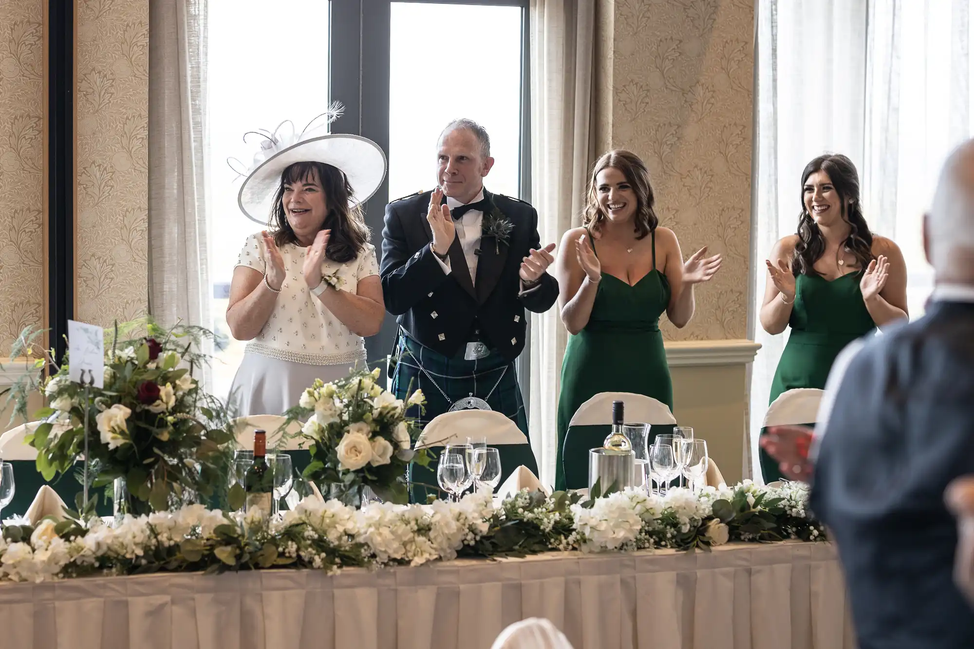 Four people stand applauding at a wedding reception, featuring two women in green dresses, a man in a suit, and a woman in a white outfit with a hat.