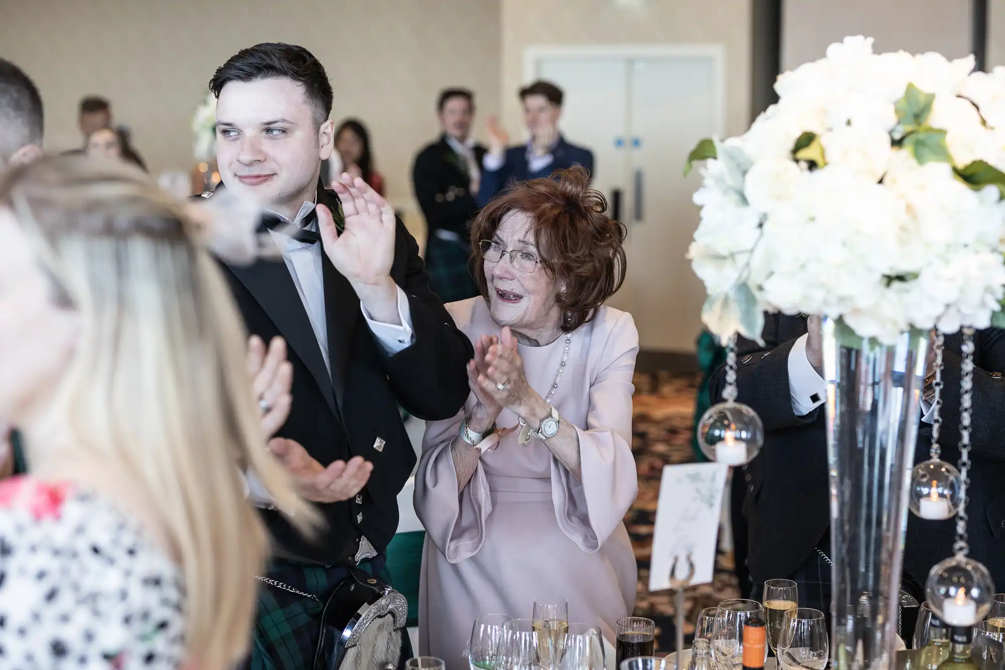 A young man in a kilt and an older woman in a lavender dress applauding at a wedding reception, surrounded by guests and floral decorations.