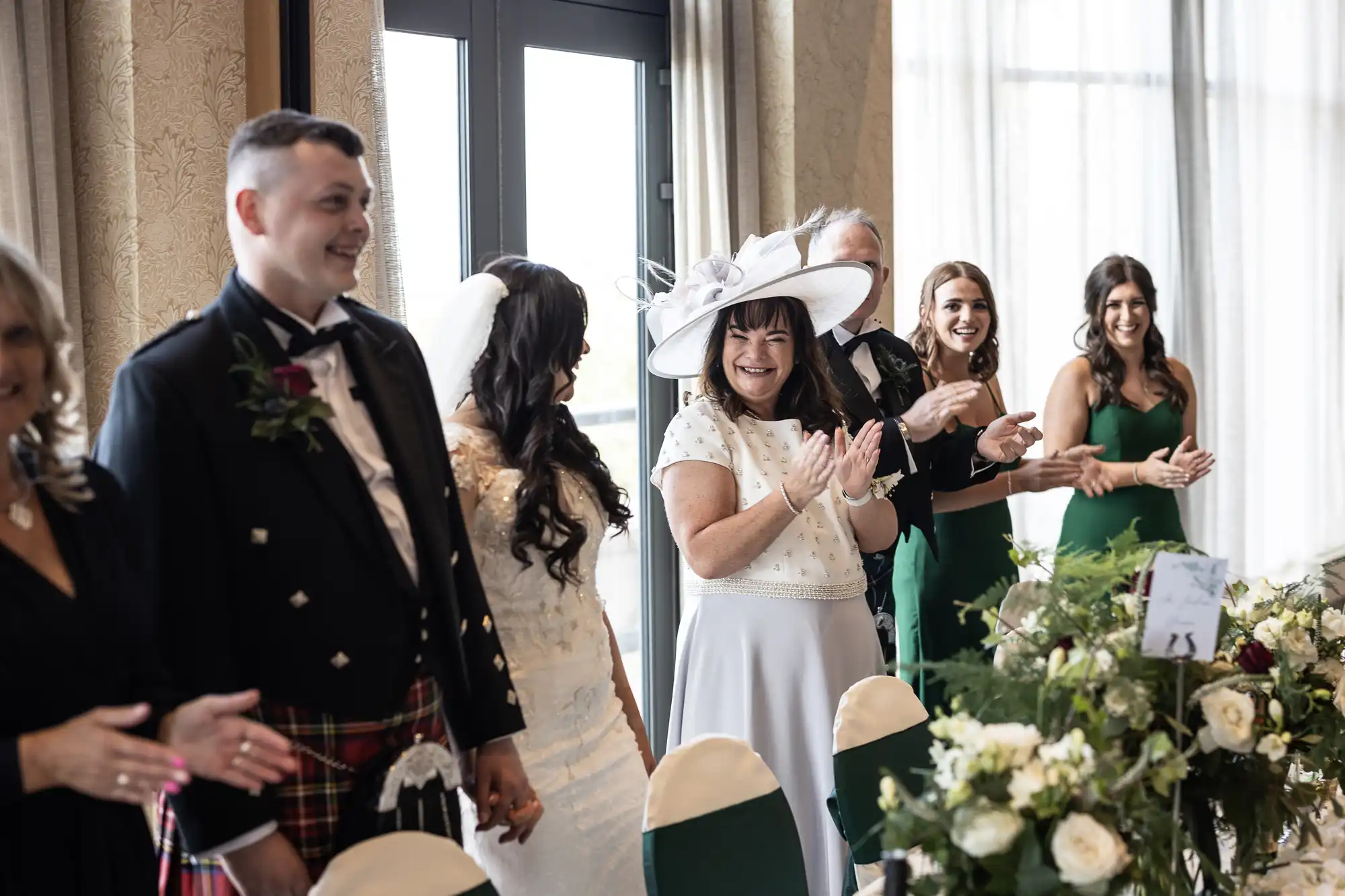 Wedding guests clapping and smiling as newlyweds walk through the venue, highlighting a mixture of formal Western and Scottish attire.