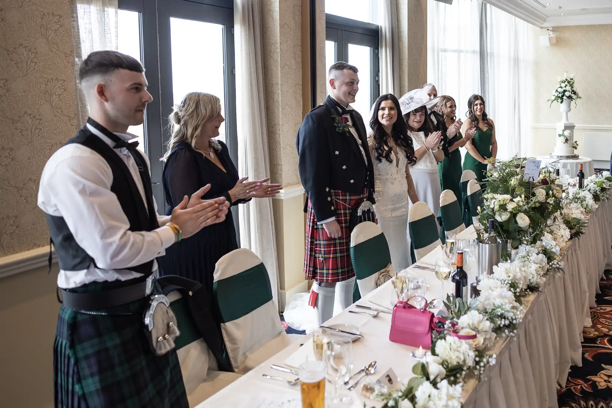 Guests in formal attire, including kilts, applaud at a wedding reception table decorated with a floral centerpiece.
