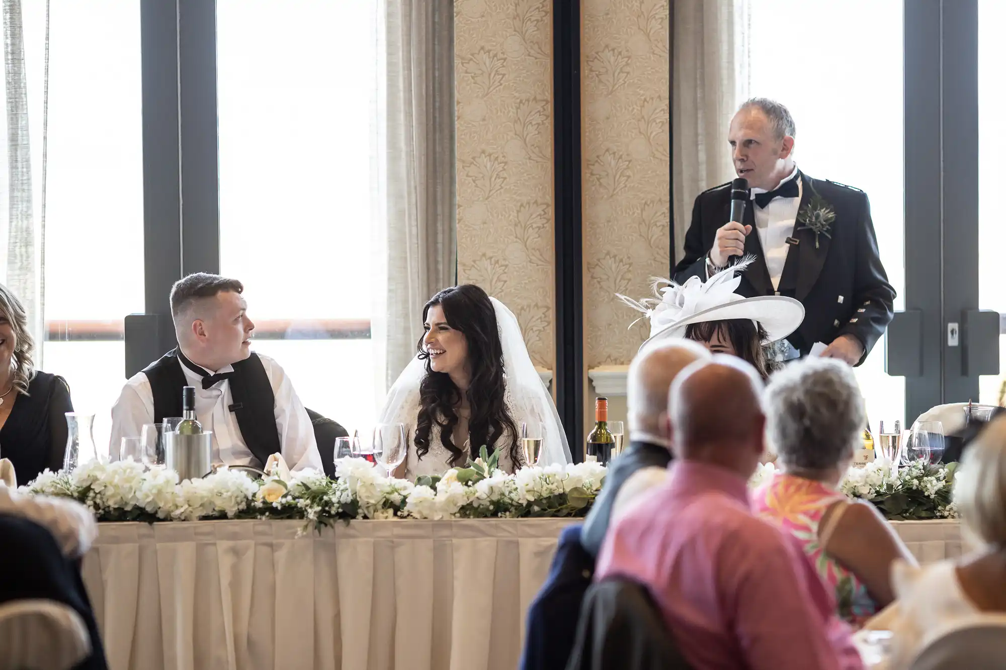 A man delivers a speech at a wedding reception, holding a microphone, with a smiling bride and groom seated at the head table, surrounded by guests and floral decorations.