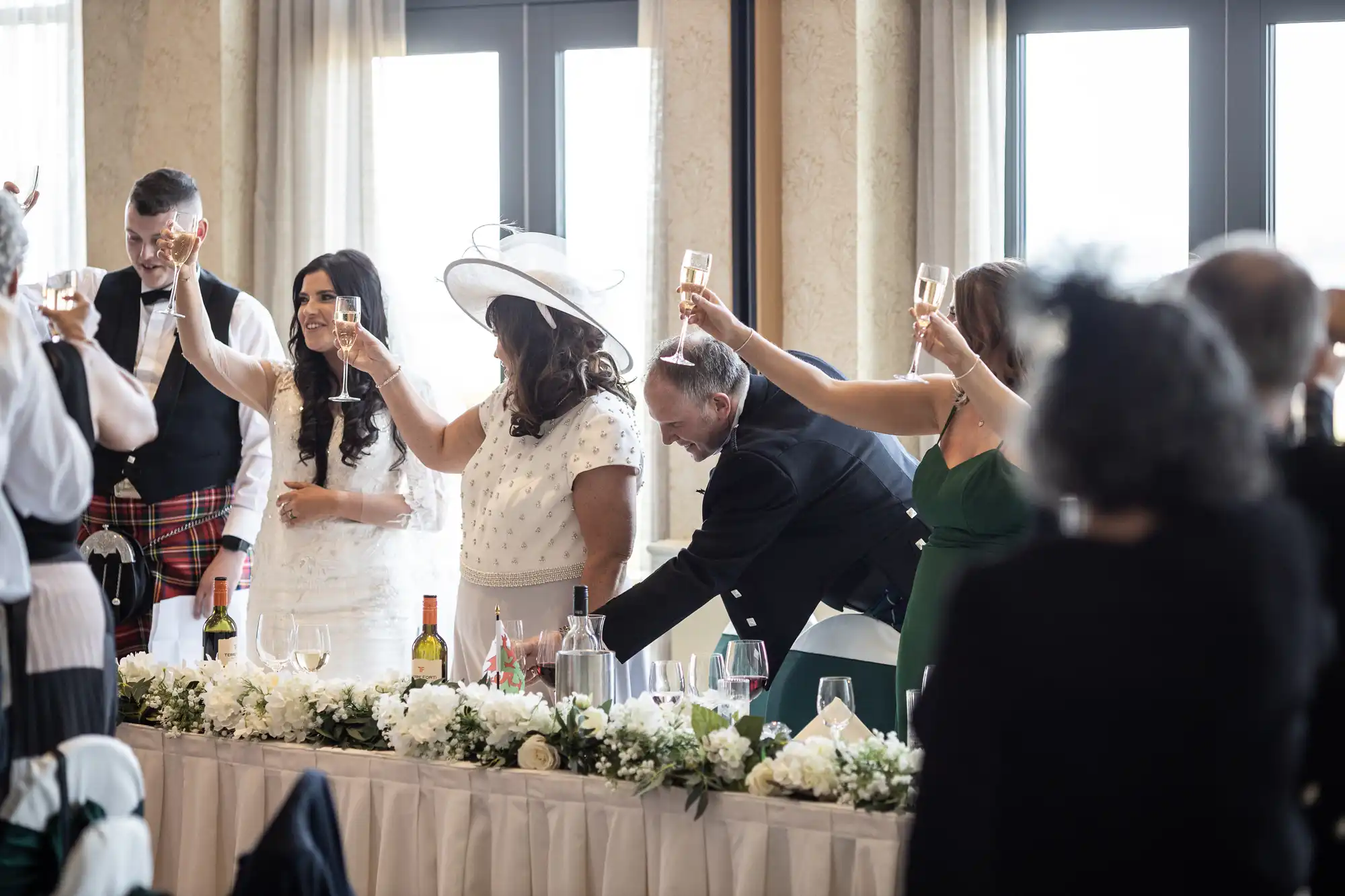 Wedding guests standing and toasting at a reception table, with a bride and groom happily raising their glasses.
