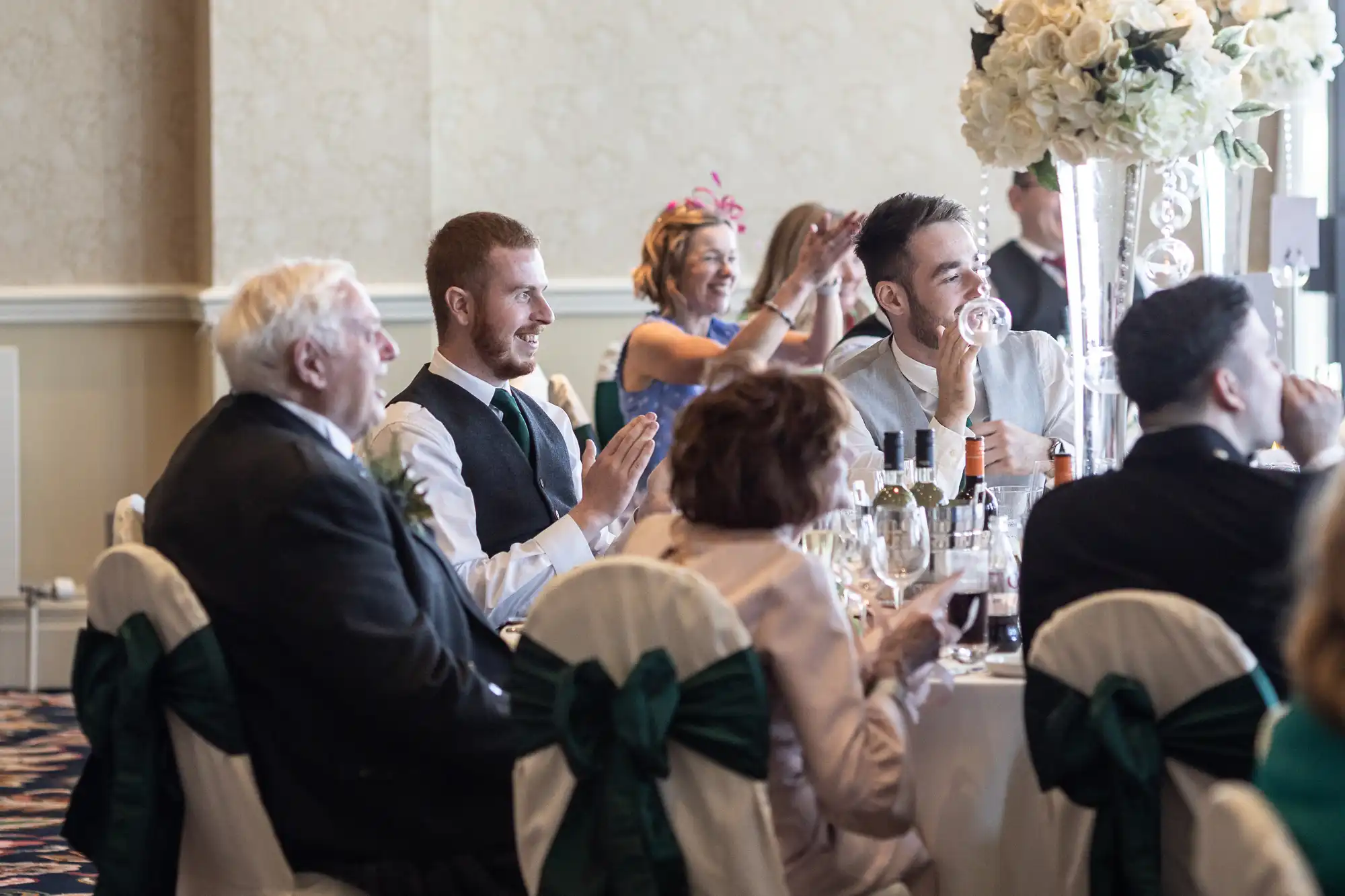 Guests at a wedding reception, clapping and smiling, seated around tables decorated with white and green floral arrangements.