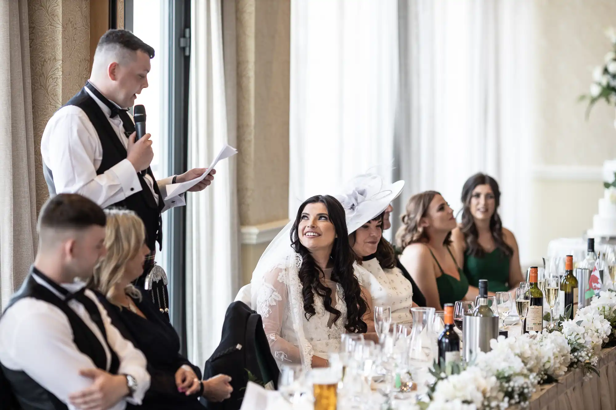 A man in a vest and bow tie gives a speech at a wedding reception, while a bride and guests, seated at a decorated table, listen attentively.