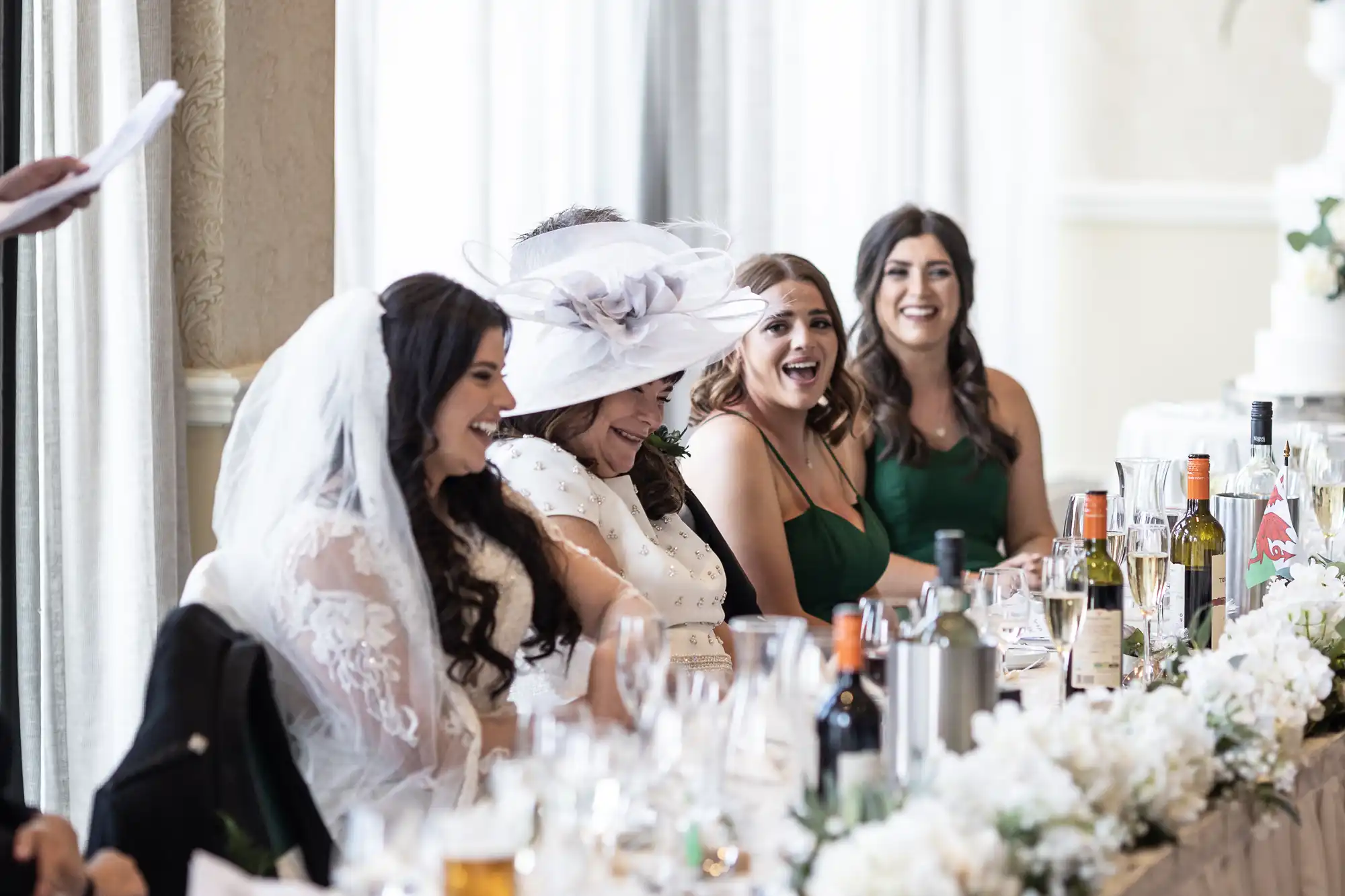 A bride in a white dress and veil laughing with three bridesmaids at a wedding reception table, surrounded by floral decorations and beverages.