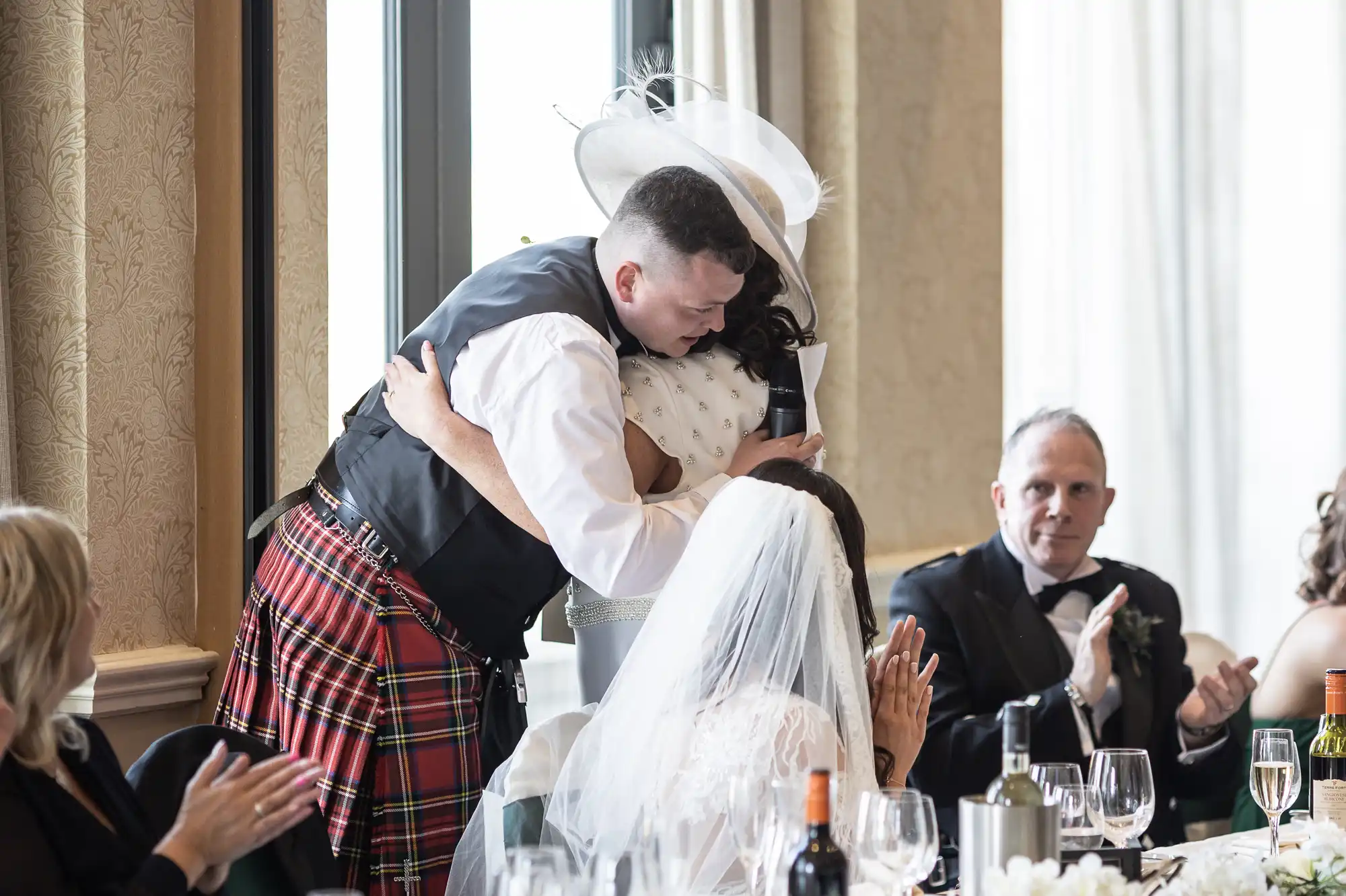 A groom in a kilt hugs a wedding guest while the bride, seated, watches; applause from other guests surrounds them in an elegant room.
