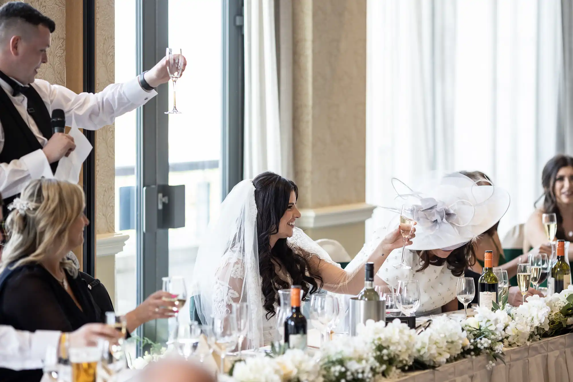A bride in a white veil and another woman in a white hat clink glasses at a wedding reception, surrounded by guests and decor.