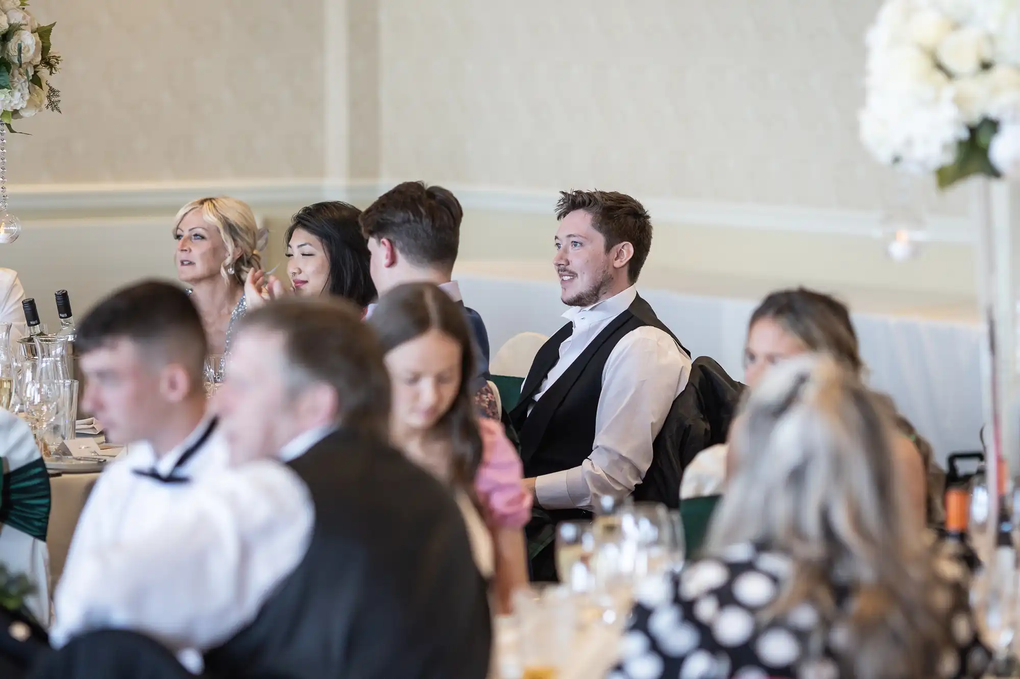 A group of guests at a wedding reception, seated at a table and engaged in conversation, with a focus on a man in a vest looking away from the camera.