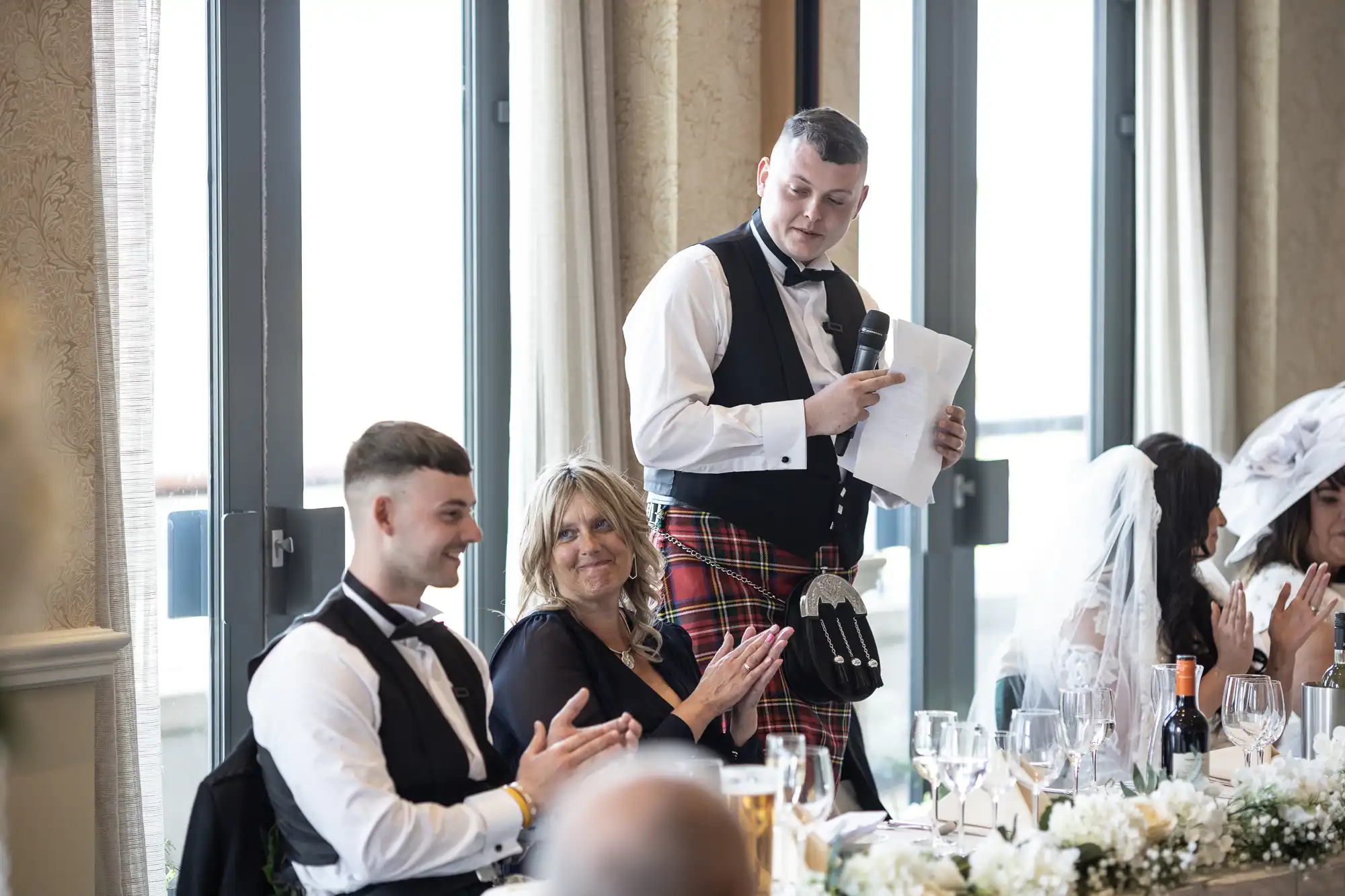 A man in a kilt and vest stands giving a speech at a wedding, holding a paper, as guests in formal attire listen and applaud in a room with large windows.