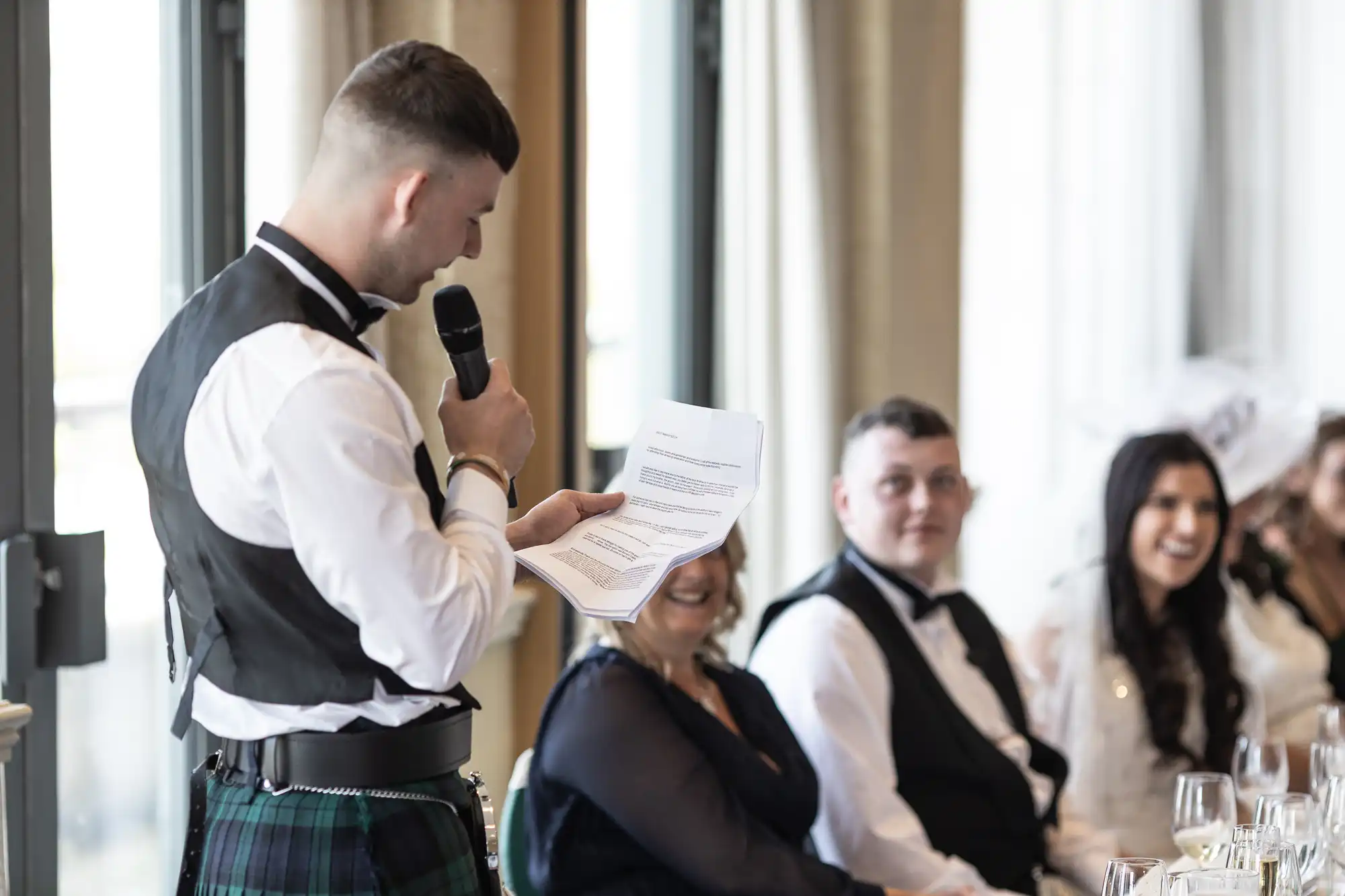 Man in kilt giving a speech with a microphone at a formal event, holding papers, with an audience of smiling people seated at a table in the background.