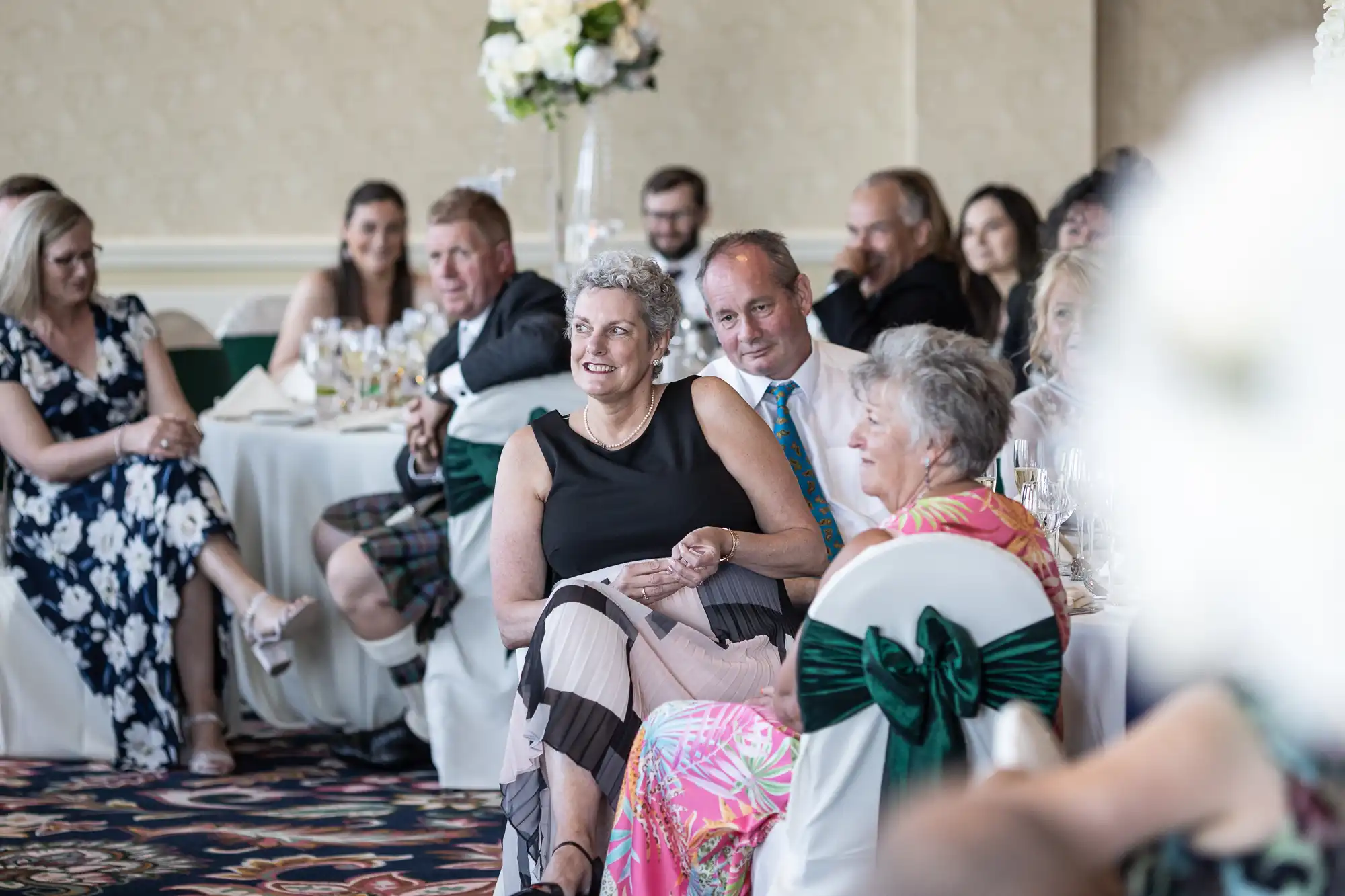 A group of elegantly dressed guests seated at a wedding reception, focused on an event happening out of frame, with a joyful expression on some faces.