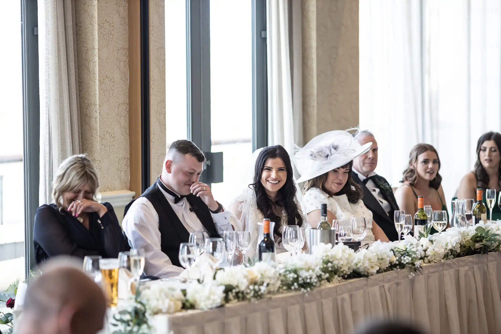 A bride and groom sit at a wedding reception table laughing with guests, surrounded by floral decorations.