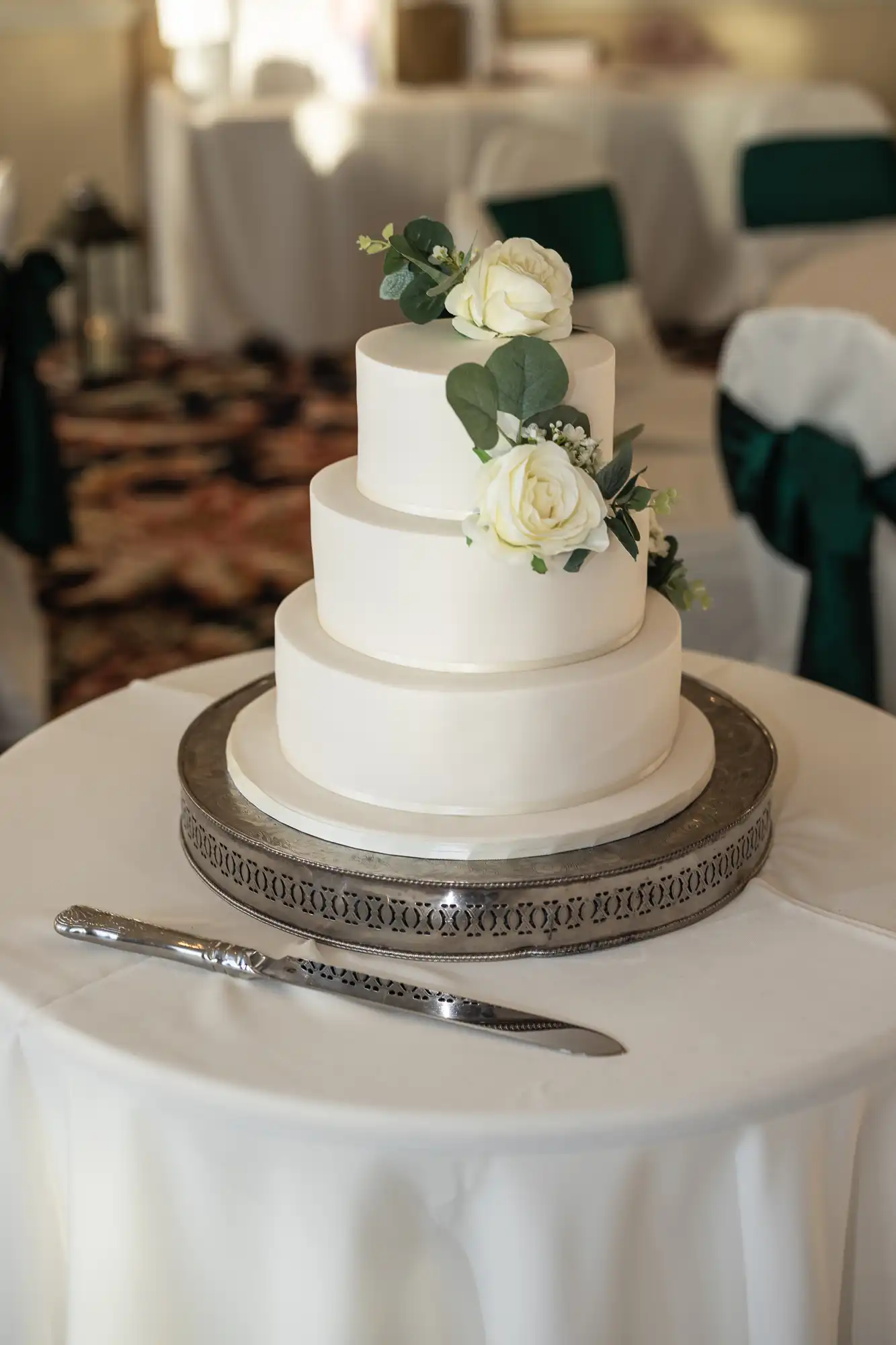 A three-tiered white wedding cake decorated with white roses on a silver stand, with a cake knife beside it, set in an elegantly decorated room.