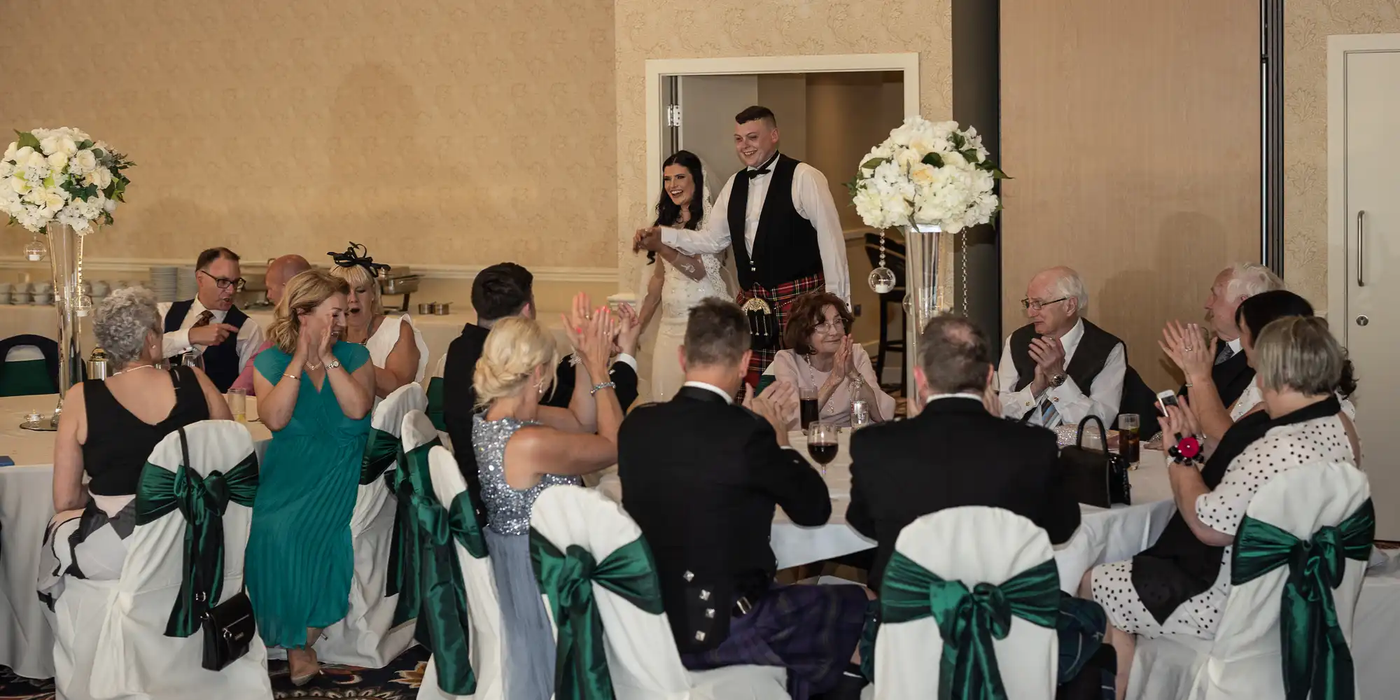 A bride and groom entering a banquet hall to applause from guests, with the groom wearing a kilt and the bride in a white dress.