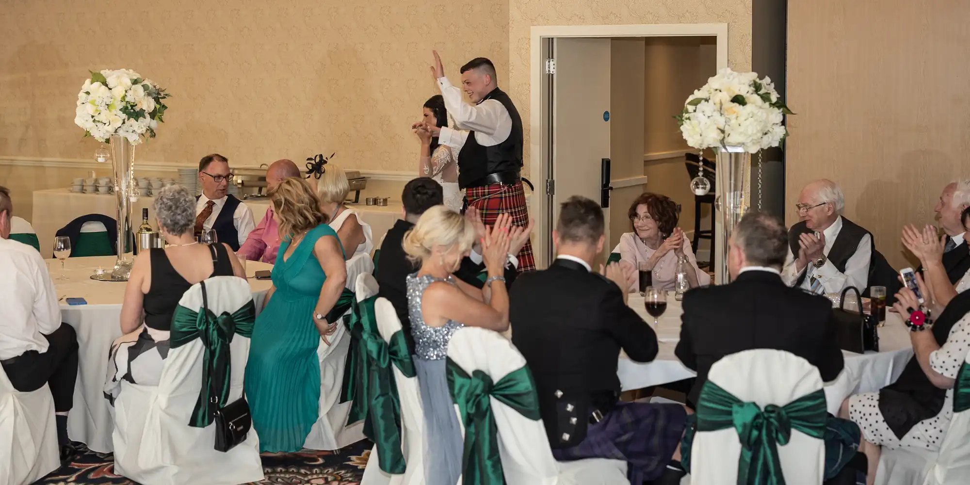 A man in a kilt is giving a speech at a wedding reception, receiving applause from guests seated at decorated tables.