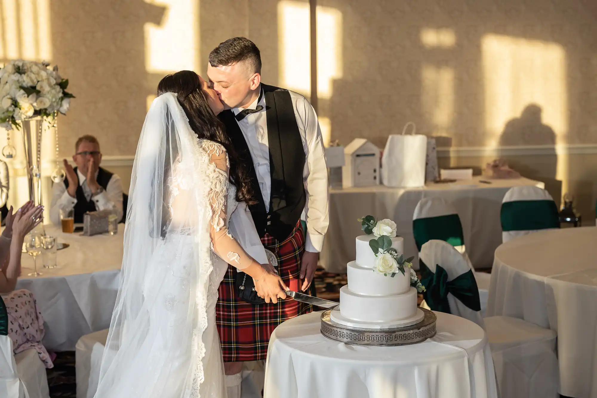 A bride and groom in a kilt cutting a wedding cake together in a sunlit room, while guests watch.