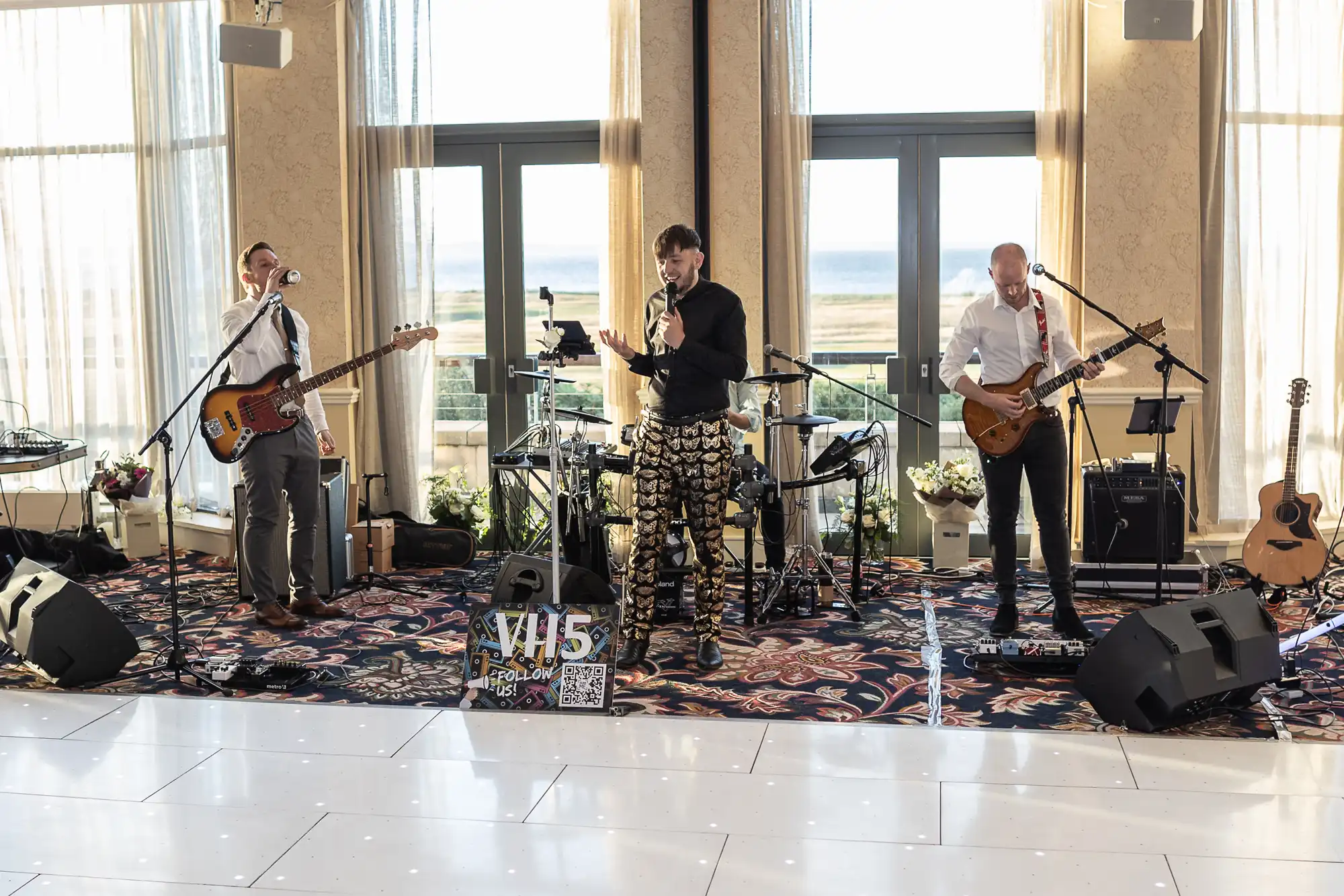 Three musicians performing indoors with ocean view, featuring a singer, and two guitarists, one also using a keyboard.