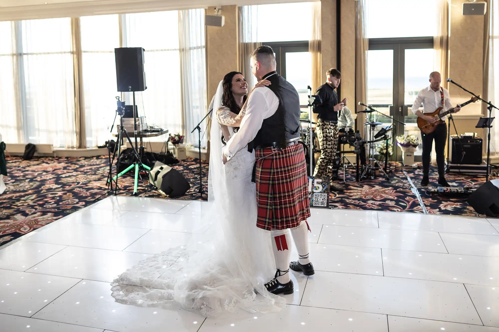 Bride in a white gown and groom in a kilt share a dance at a wedding reception, with a live band performing in the background.