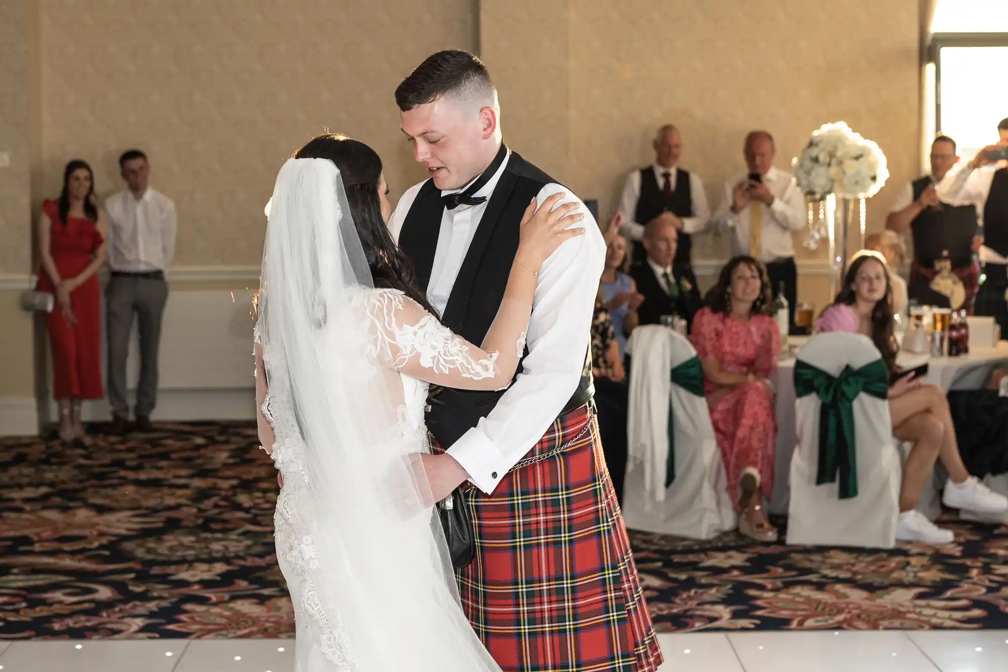 A bride and groom share their first dance at their wedding reception, surrounded by guests, with the groom wearing a kilt.