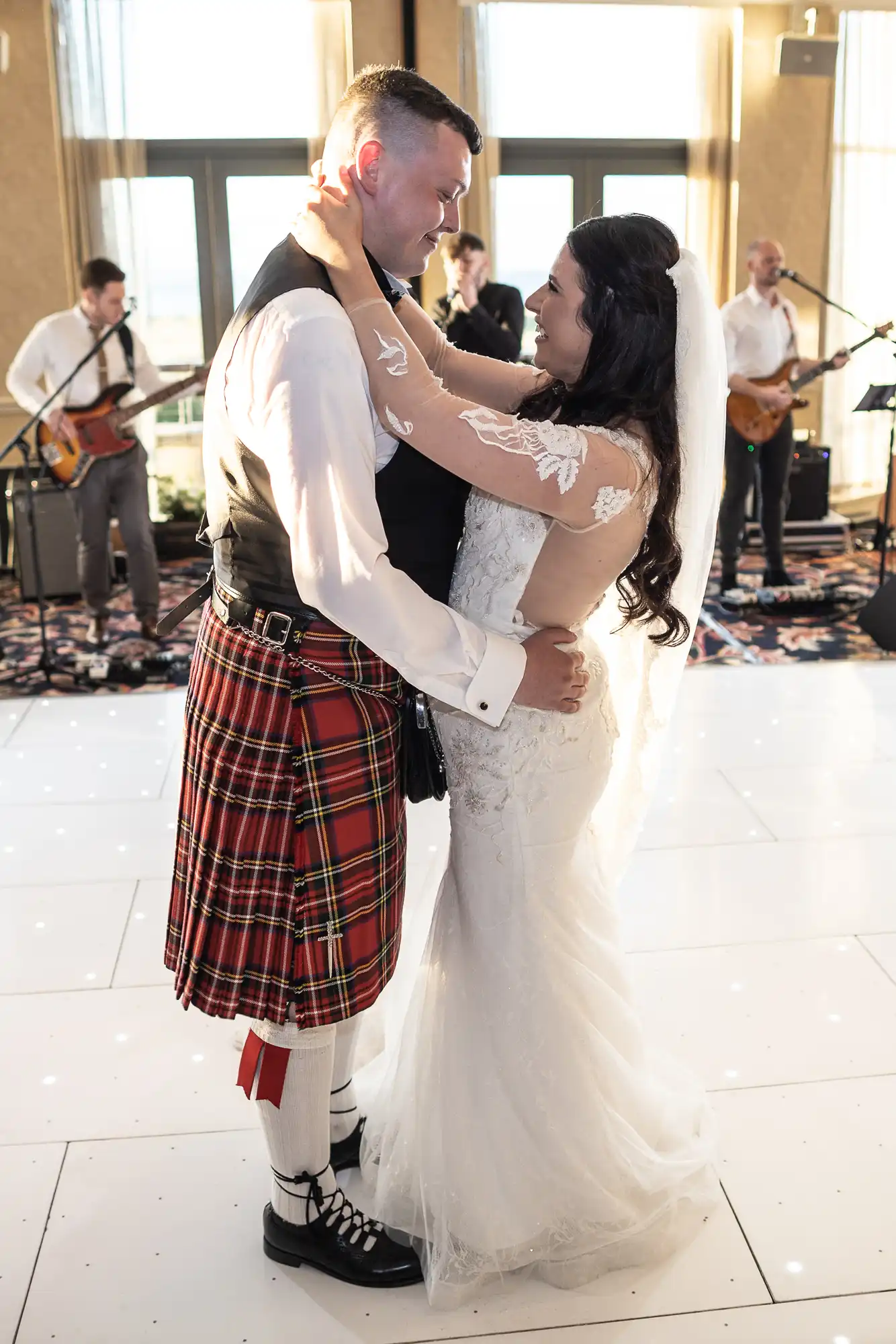 A bride in a white lace dress and a groom in a tartan kilt share a joyful dance at their wedding reception, with a band performing in the background.