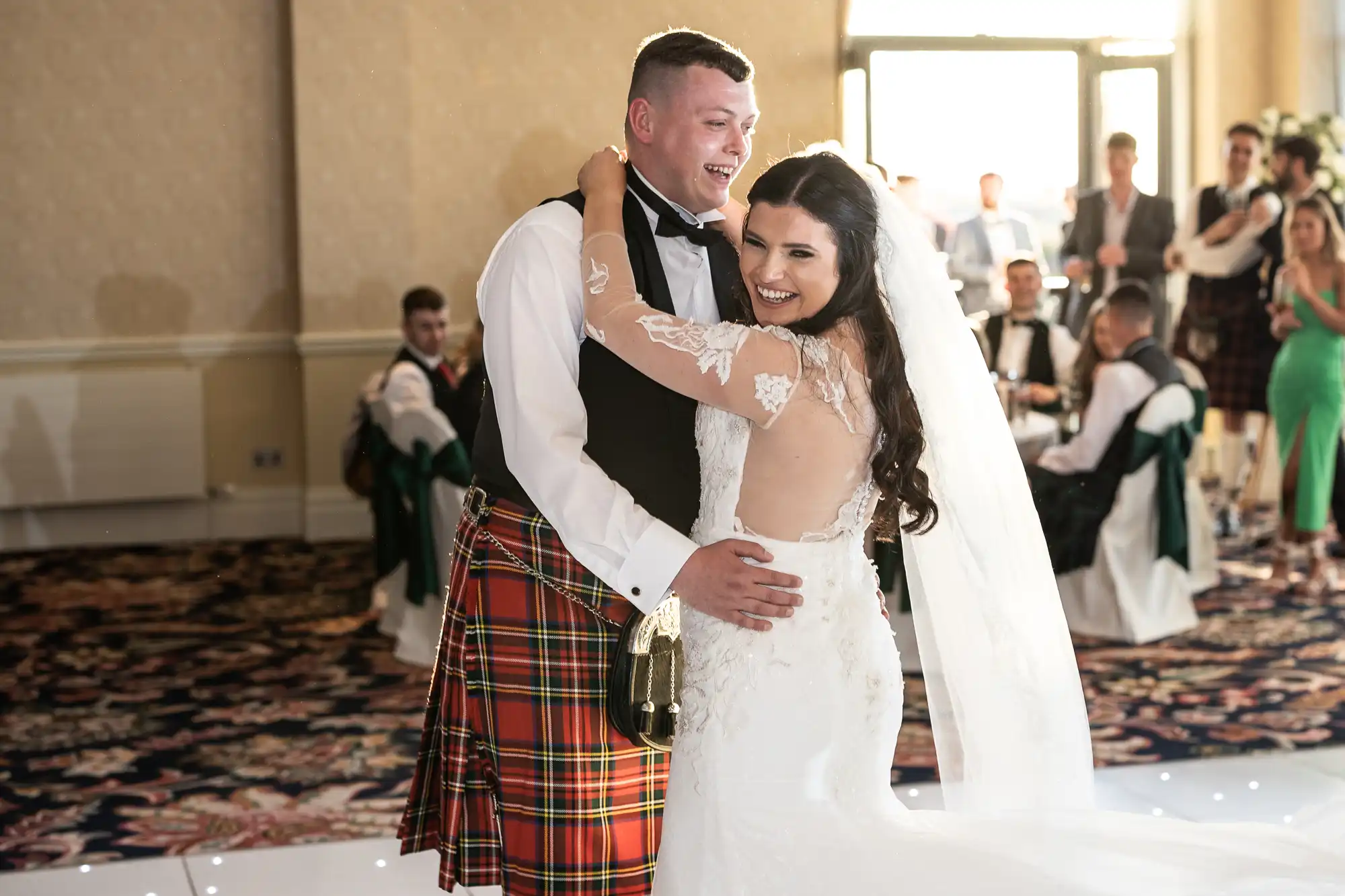 A bride and groom smile and embrace on the dance floor, surrounded by guests. The groom wears a tartan kilt, and the bride is in a lace gown.