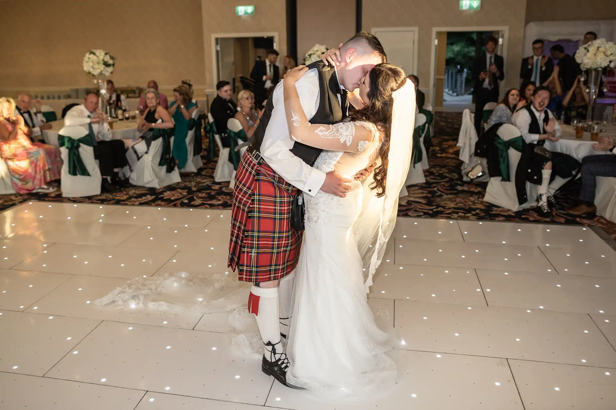 A bride and groom embracing and kissing on a dance floor, the groom wearing a kilt, with wedding guests watching in the background.