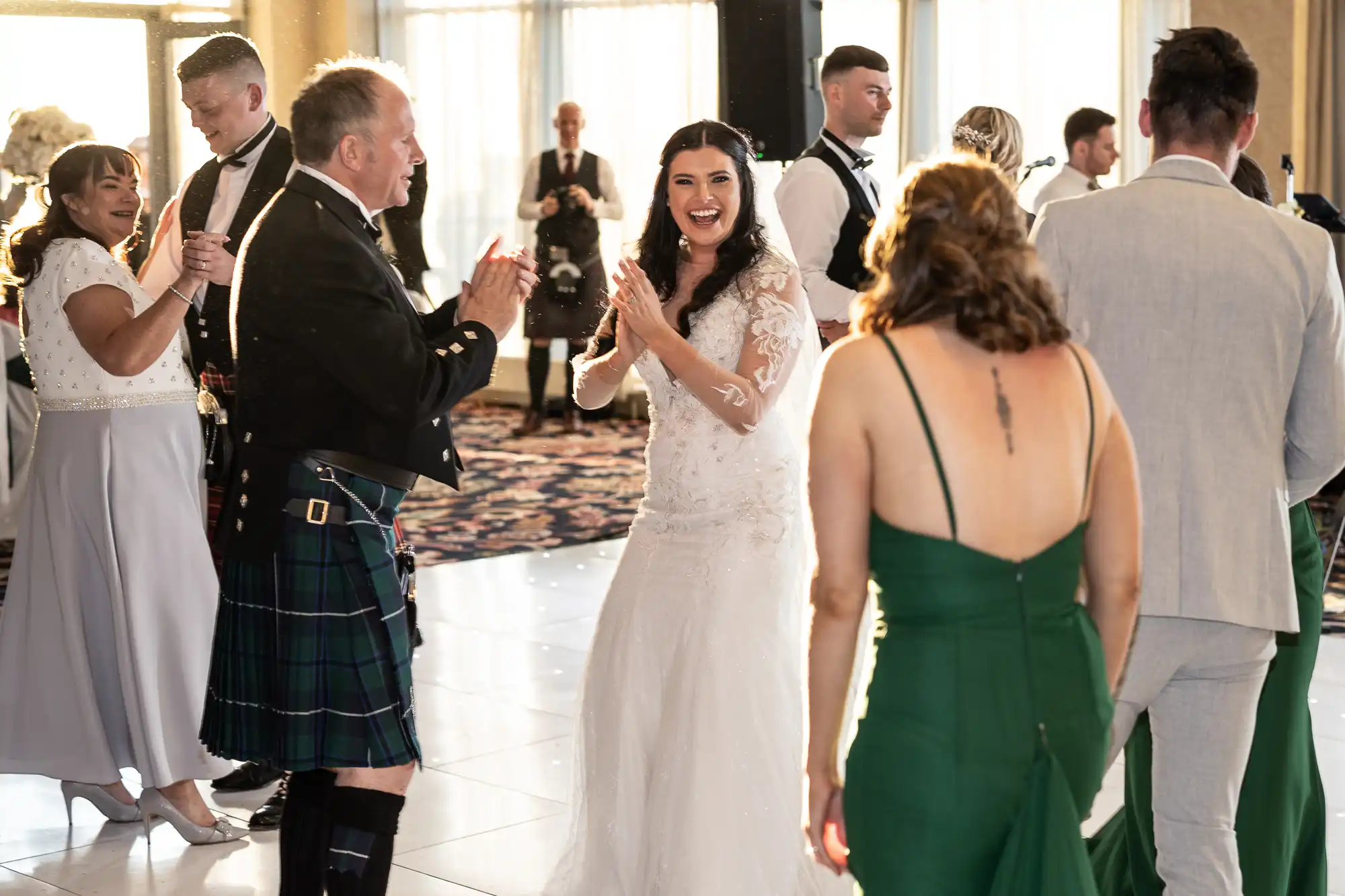 A bride in a lace dress joyfully claps hands with a man in a kilt at a lively wedding reception, surrounded by guests in formal attire.