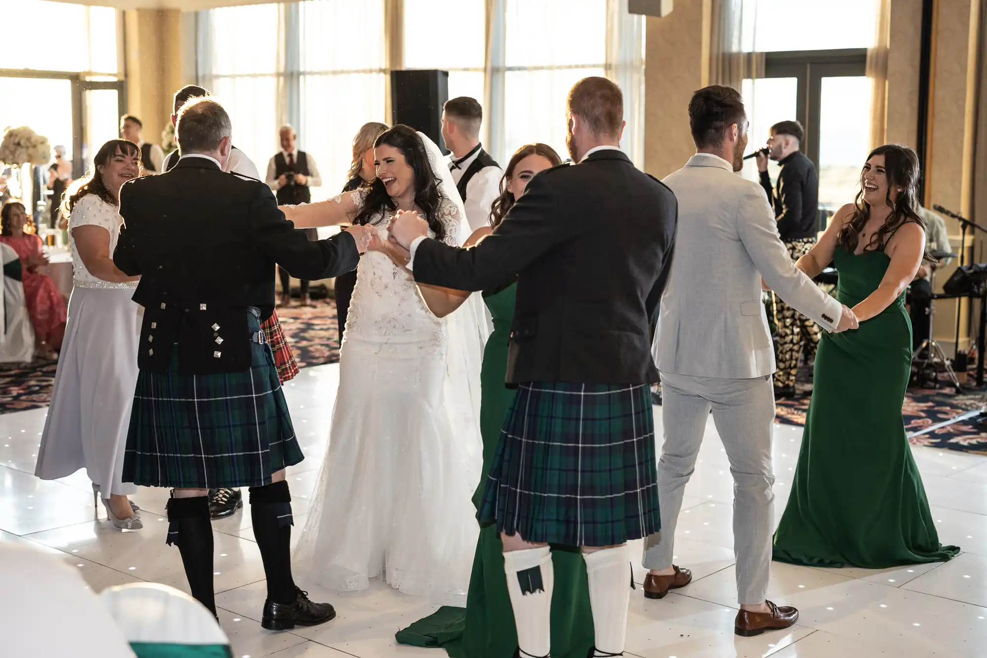 A bride and groom joyfully dance surrounded by guests, some wearing Scottish kilts, in an elegant hall with large windows.