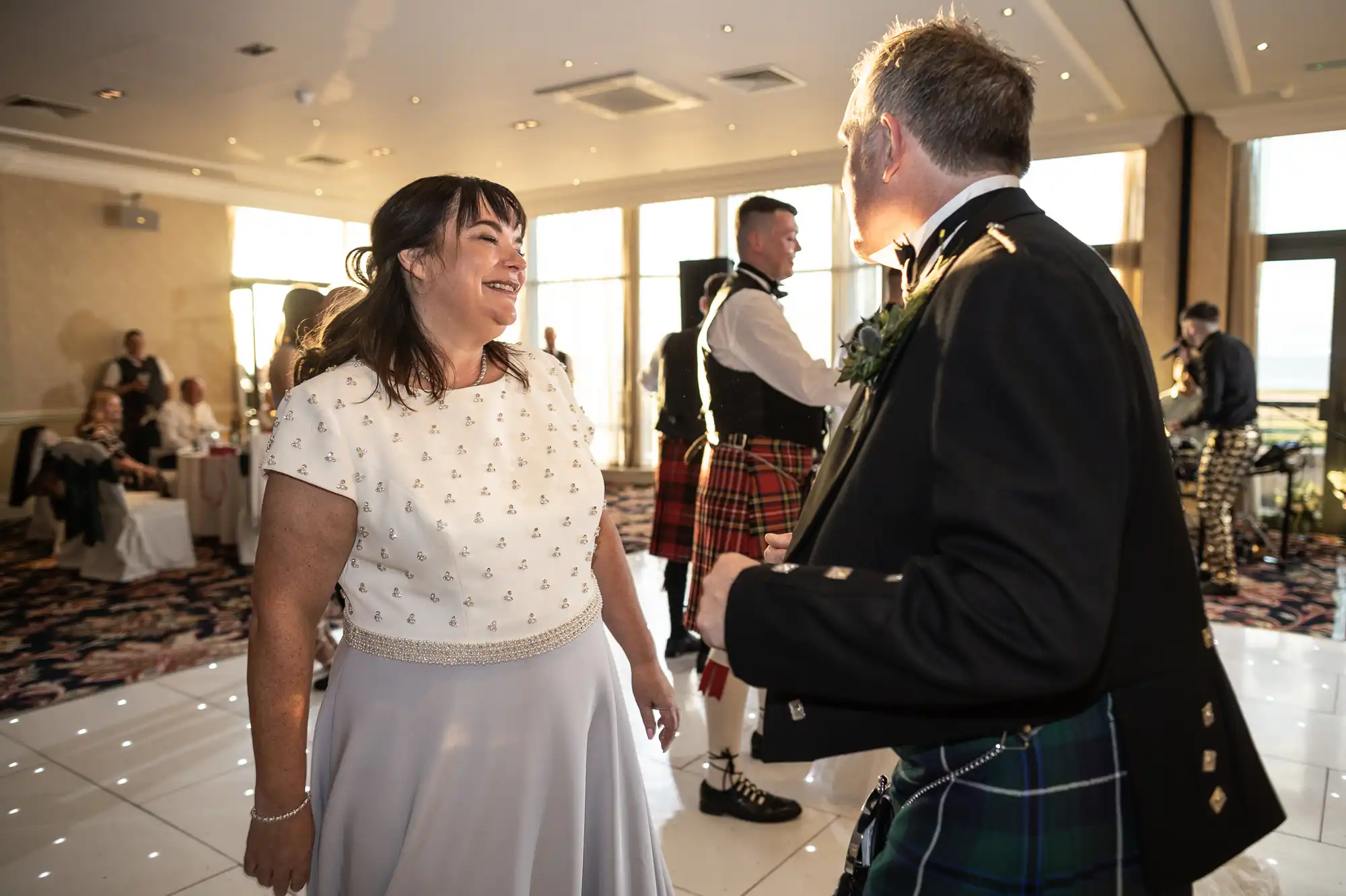 A woman in a white dress smiles widely at a man in a traditional Scottish kilt during an indoor event with bagpipers in the background.