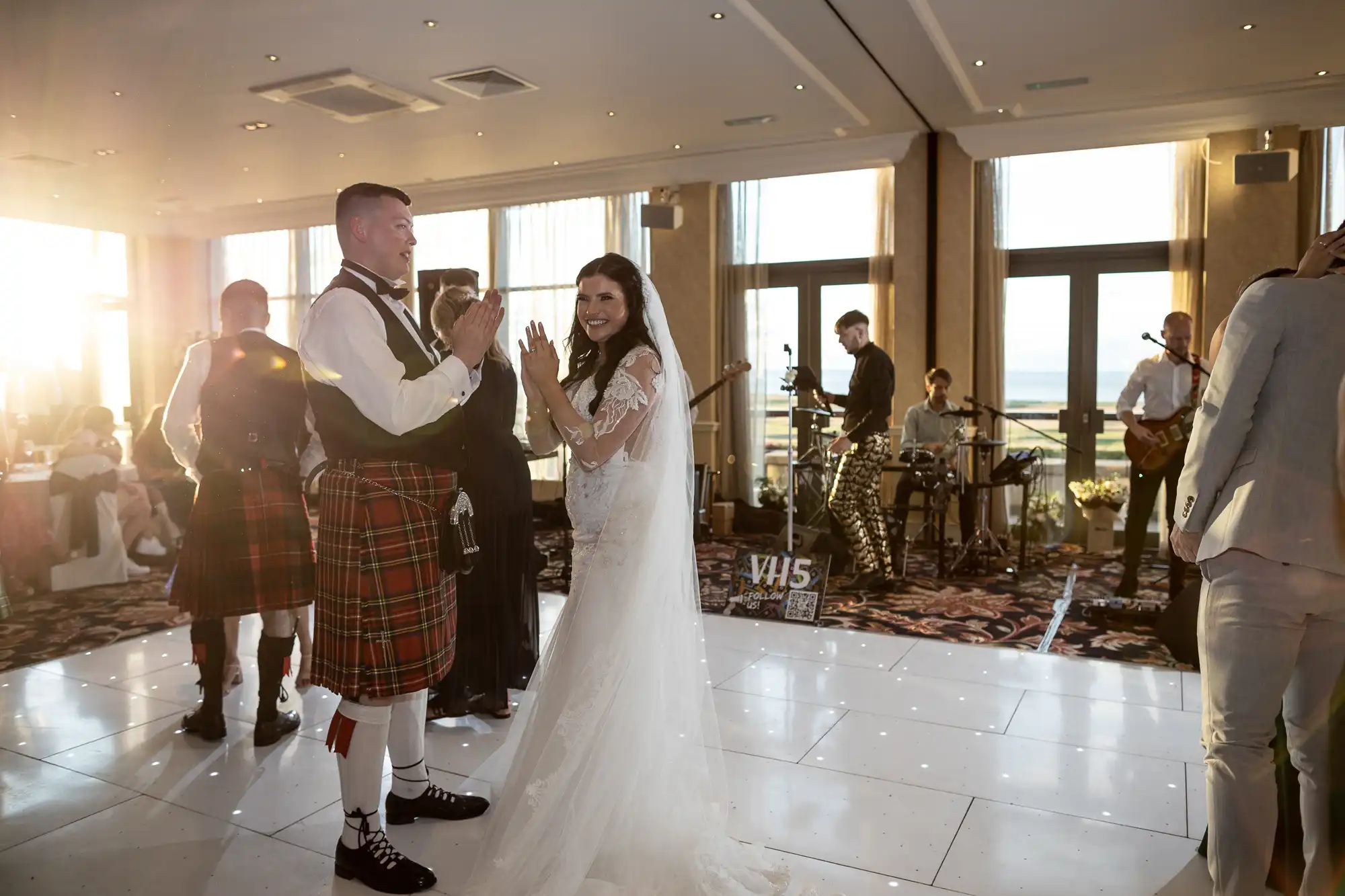 A bride and groom in a joyful dance at a wedding reception, the groom wearing a traditional kilt, with guests and a live band in the background.