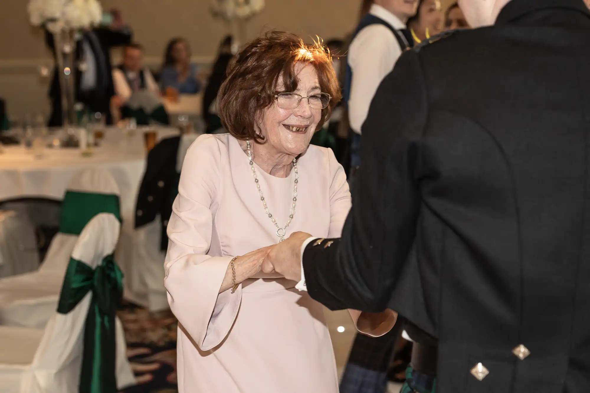 Elderly woman in a pink dress and glasses smiles while shaking hands with a man in a dark suit at a festive event.