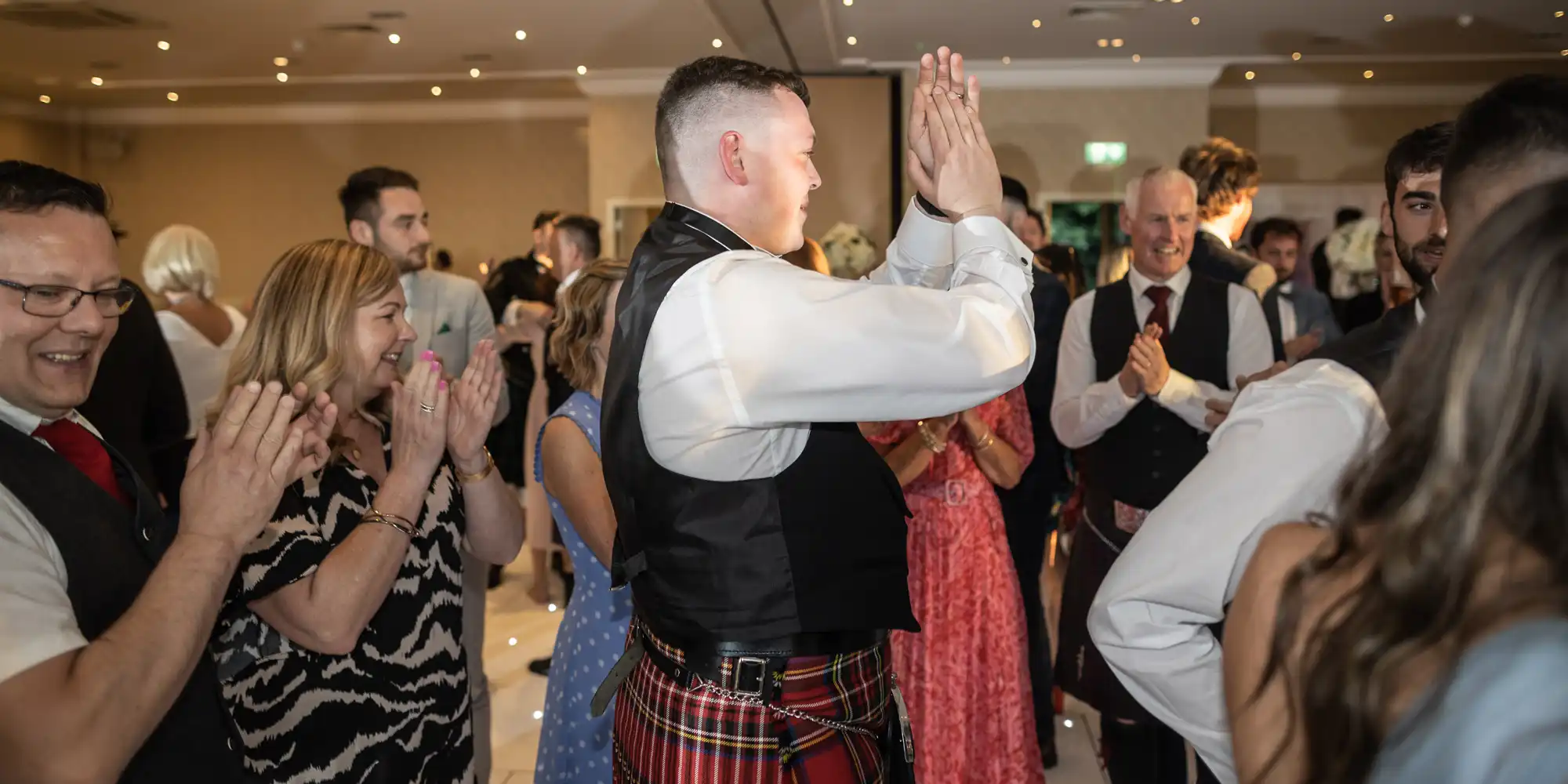 A man in a kilt high-fives another man as people around them clap and celebrate at an indoor event.